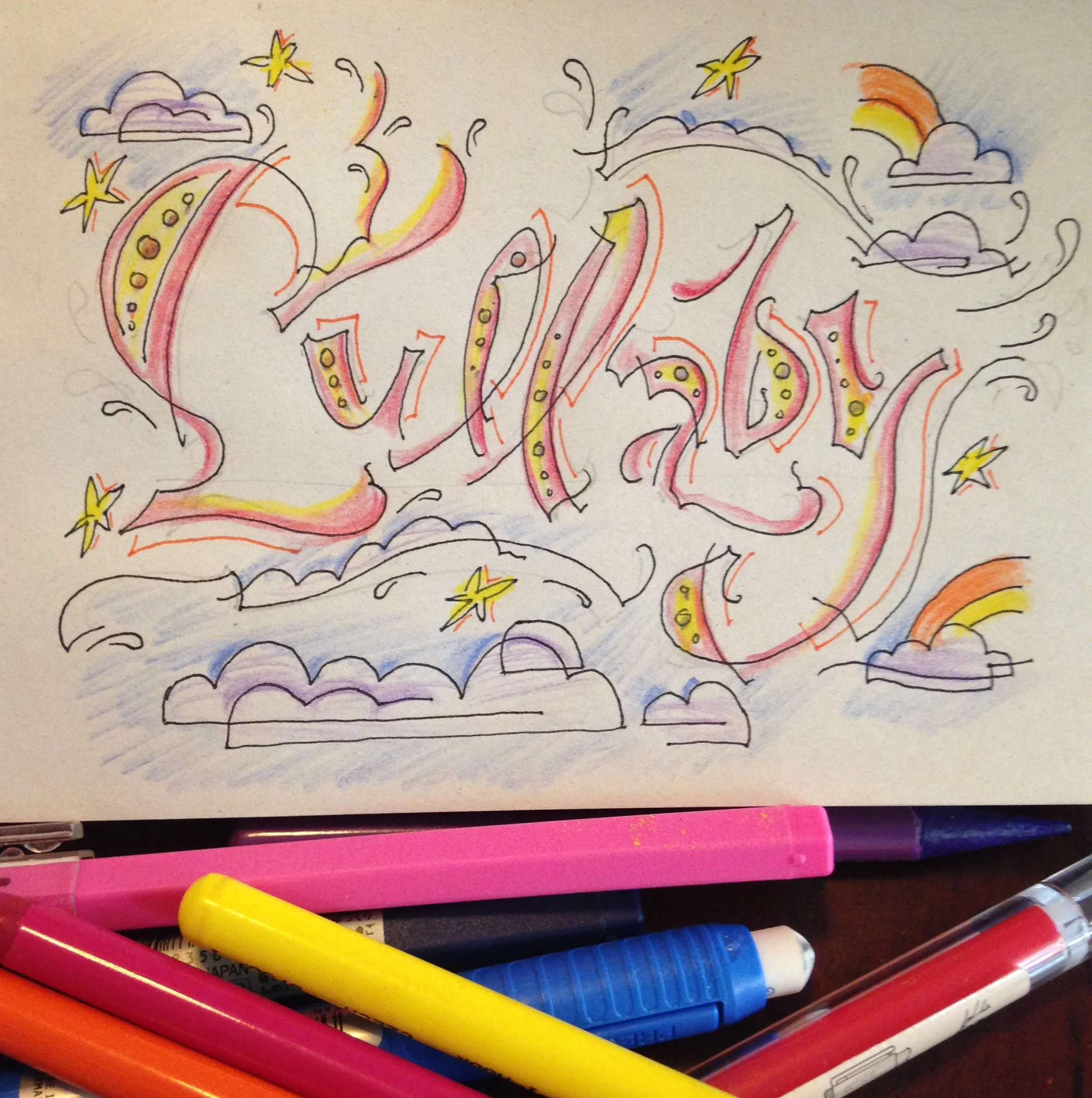 Hand-drawn lettering of the word "lullaby" with colorful decorative elements such as stars, clouds, and rainbows, surrounded by pens and markers.