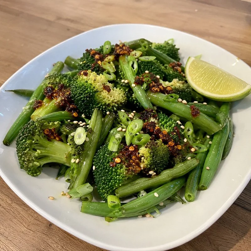 Healthy steamed vegetables with chili crisp, broccoli, green beans, sesame seeds, green onion, and lime on a plate.