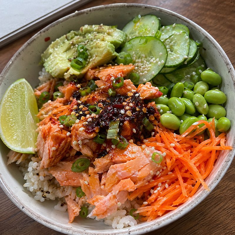 Healthy chili crisp salmon rice bowl with flaky salmon, brown rice, cucumber, edamame, avocado, green onion, sesame seeds, and spicy chili crisp.
