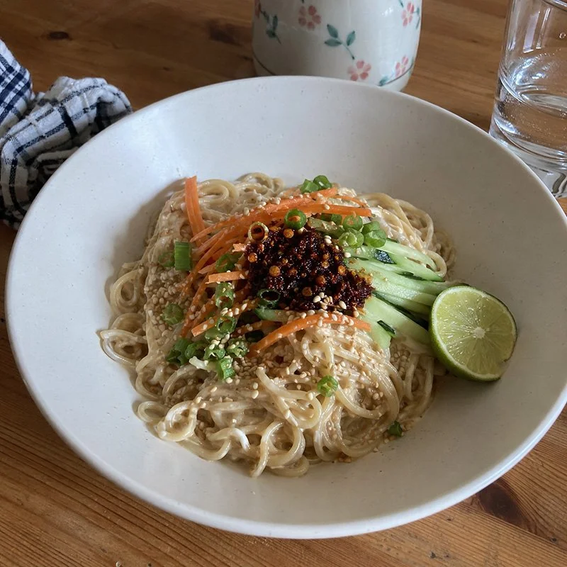 Yuzu-sesame cold noodles with chili crisp, cucumber, carrots, green onion, sesame seeds, herbs, and citrusy sesame sauce in a bowl.