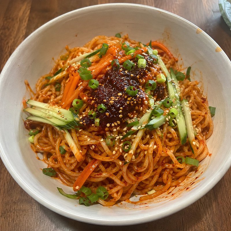 Spicy gochujang noodles with chili crisp, cucumber, carrots, green onion, sesame seeds, and chili oil in a bowl.