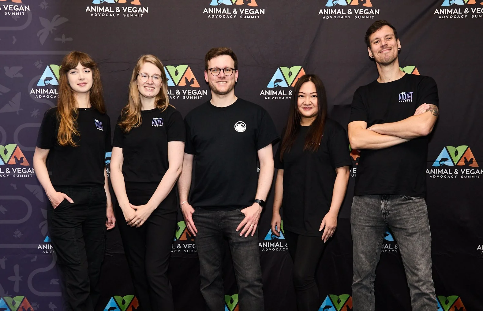 Group of five people standing in front of an Animal & Vegan Advocacy Summit backdrop, wearing black shirts with event logos, smiling and posing for the photo.