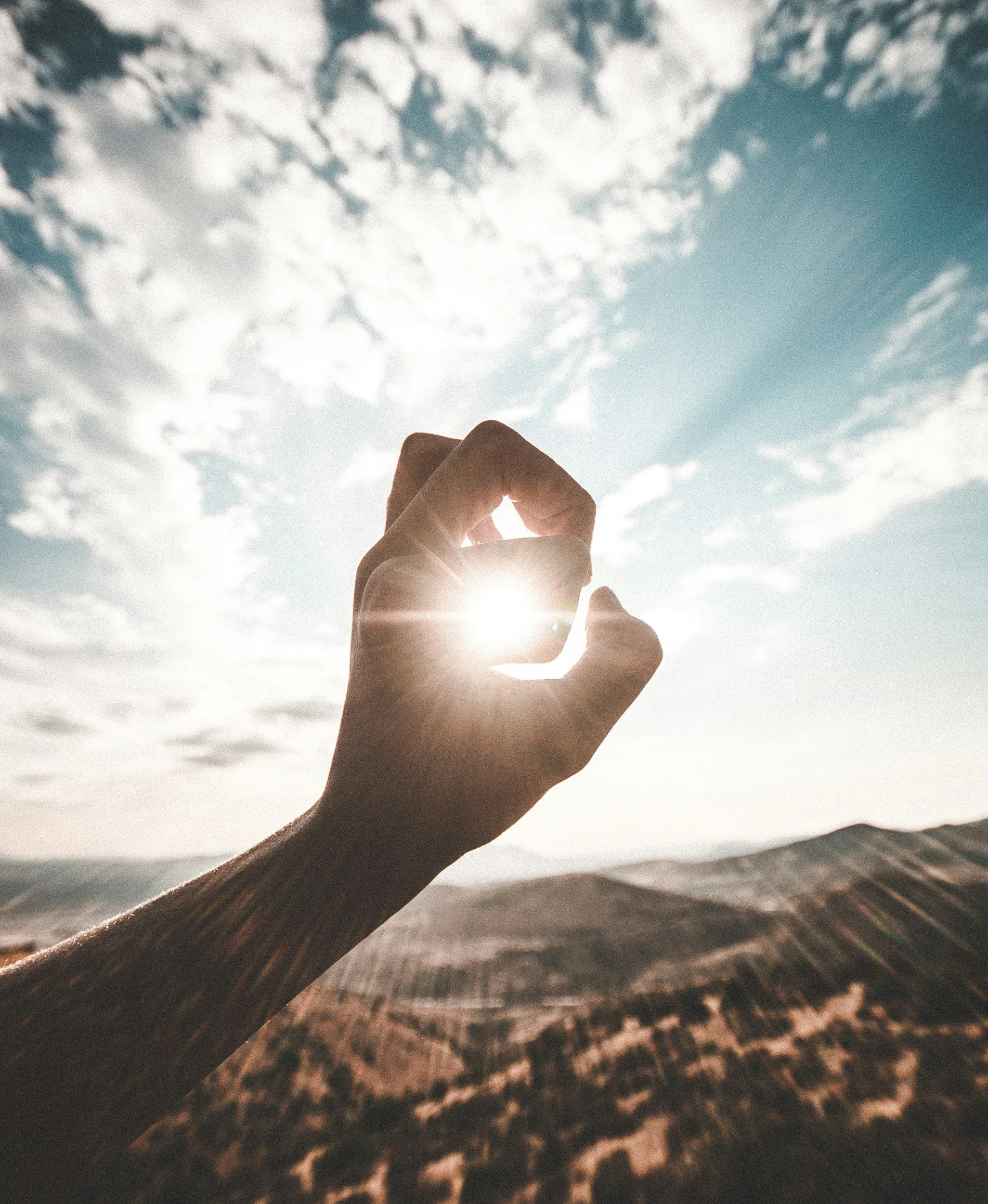 A hand making an 'OK' gesture with the thumb and index finger forming a circle, held up against a bright sky with scattered clouds, during sunset or sunrise, with a landscape of hills or mountains in the background.
