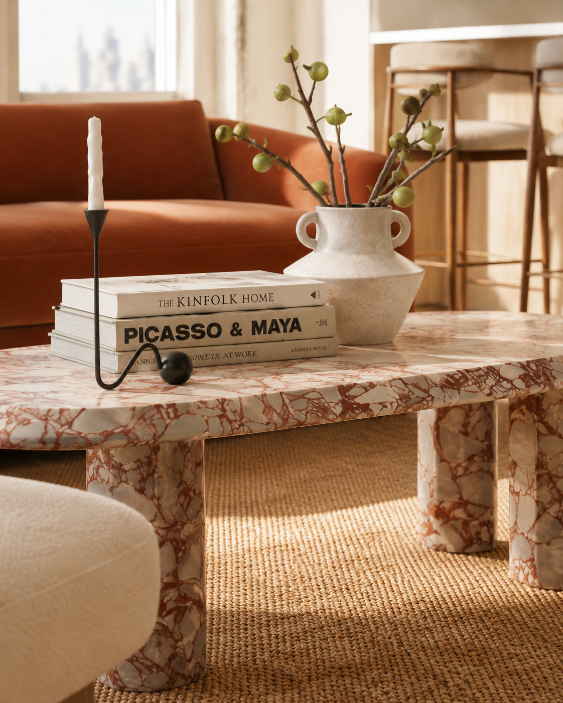 A marble coffee table with a stack of books titled 'The Kinfolk Home', 'Picasso & Maya', and another book. A white vase with green branches is on the table, along with a black and white modern candle holder. In the background, a rust-colored sofa and bar stools are visible in a cozy living room.