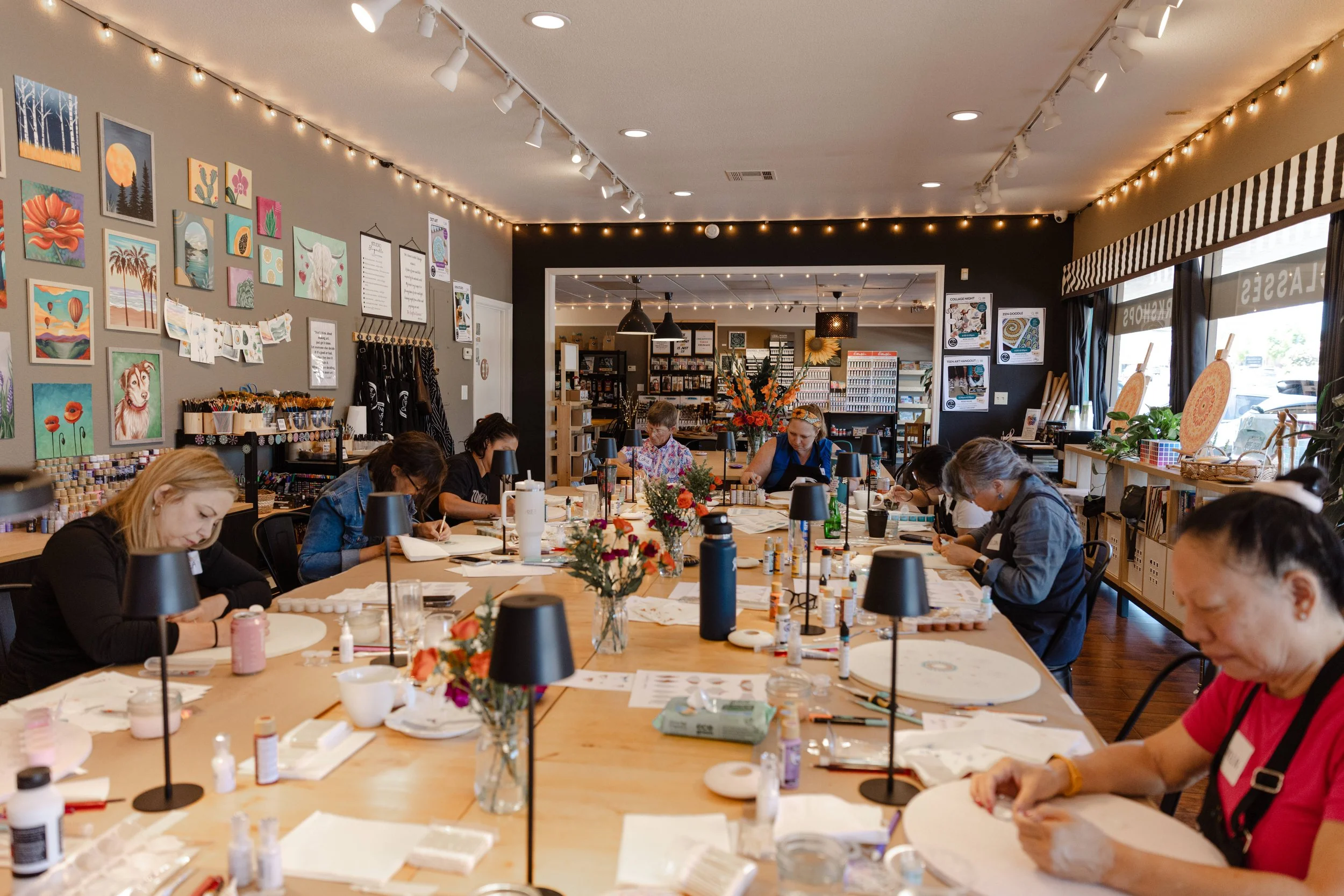 People participating in an art or craft workshop inside a well-lit, decorated room with artwork on the walls, flowers on the table, and shelves with art supplies.