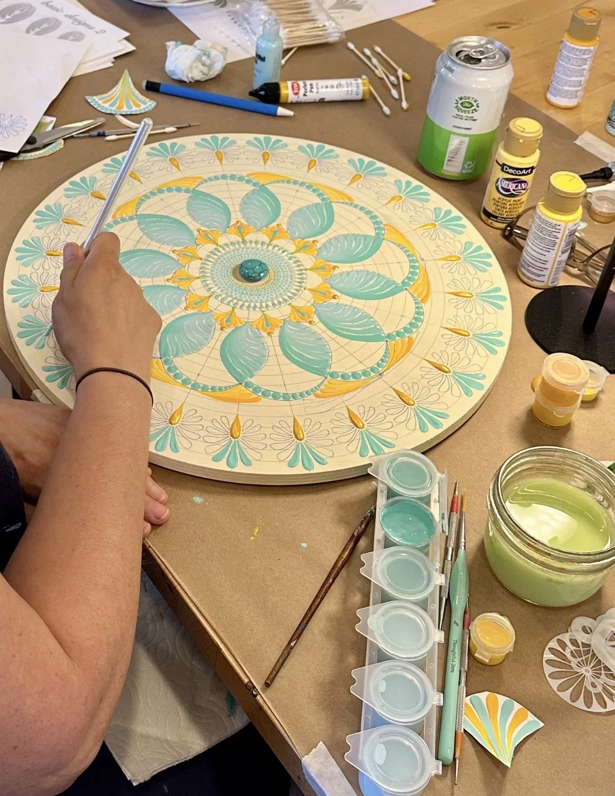 A person creating a colorful mandala design on a round wooden art piece using teal, yellow, and white paints, with art supplies and water containers surrounding the workspace.