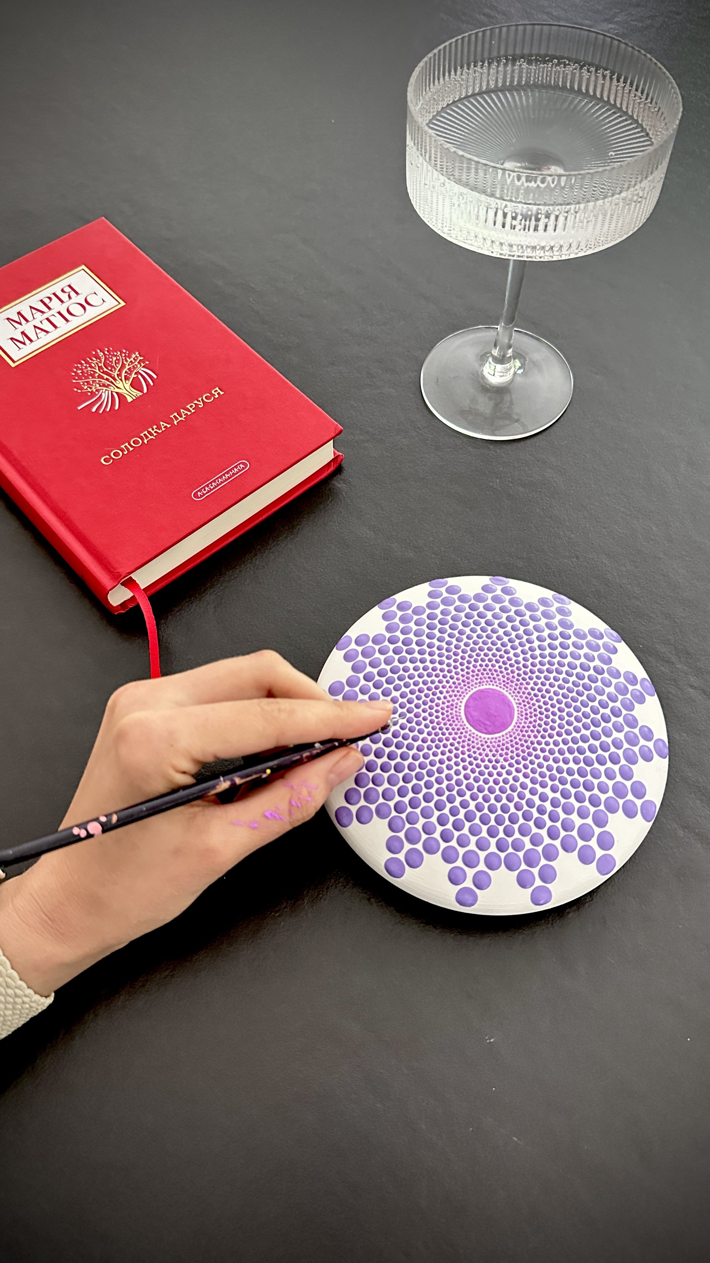 A person holds a paintbrush applying pink dots to a circular mandala design on a white canvas. On the table, there is a red Russian book with gold lettering, a clear glass, and an optical illusion artwork with purple dots and concentric patterns.