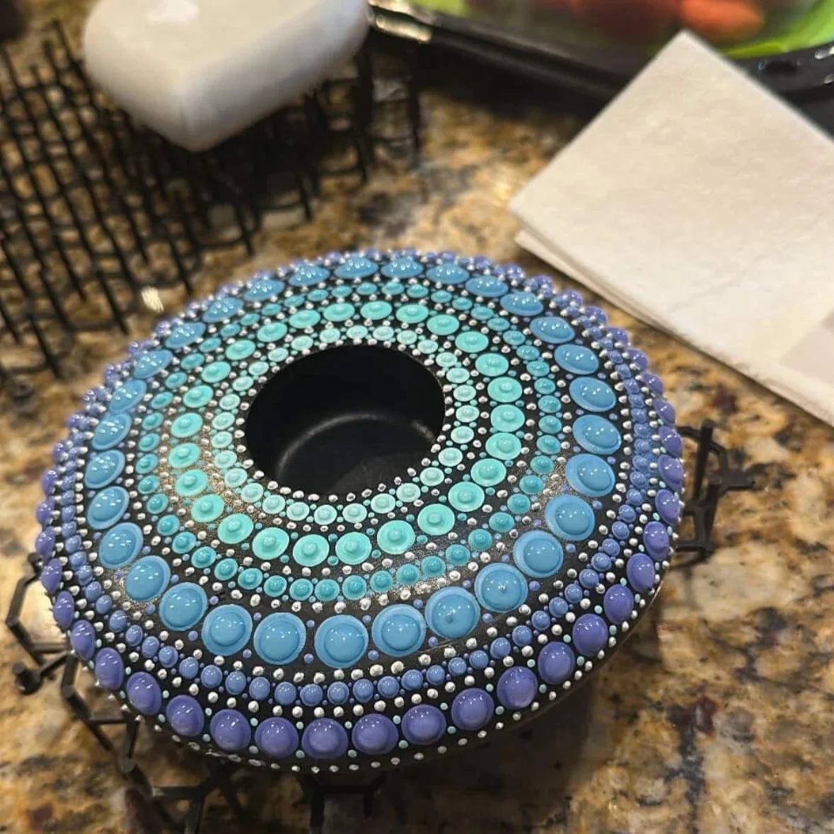 Decorative black candle holder with intricate blue and purple bead designs arranged in concentric circles, placed on a granite kitchen countertop.