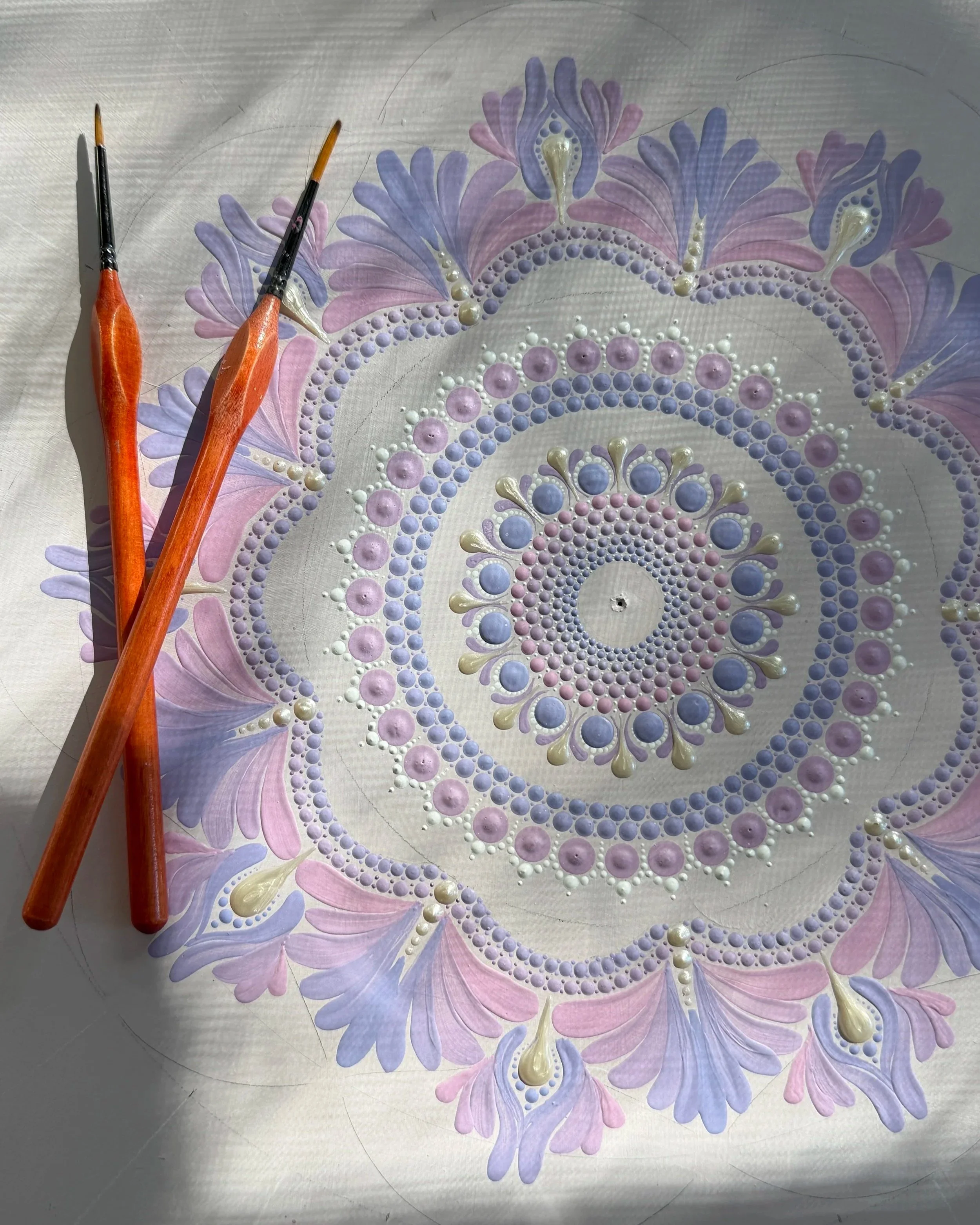 A decorative artwork featuring pastel-colored floral patterns with intricate bead and pearl detailing, on a metallic surface. Two paintbrushes with orange handles are resting on the artwork.