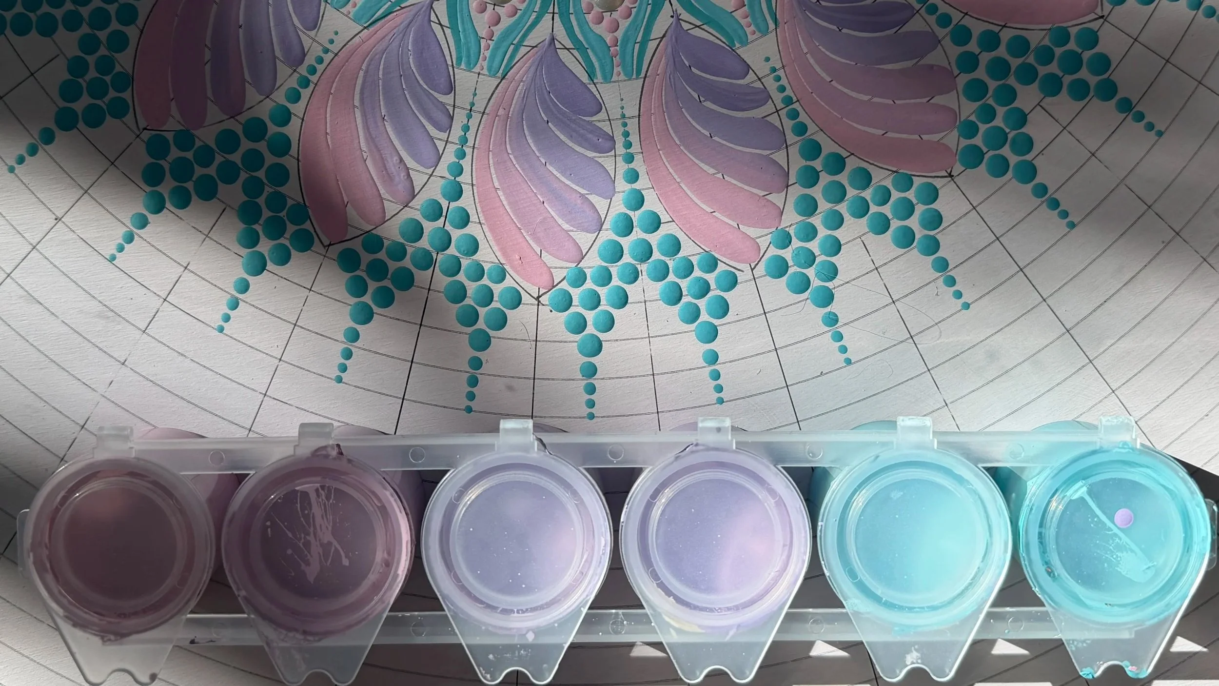 Close-up of a watercolor painting or art project with colorful leaf designs in pastel shades of purple, pink, and teal, along with a pattern of teal dots, on grid-lined paper. There are also small containers of paint or watercolor pigments at the bot