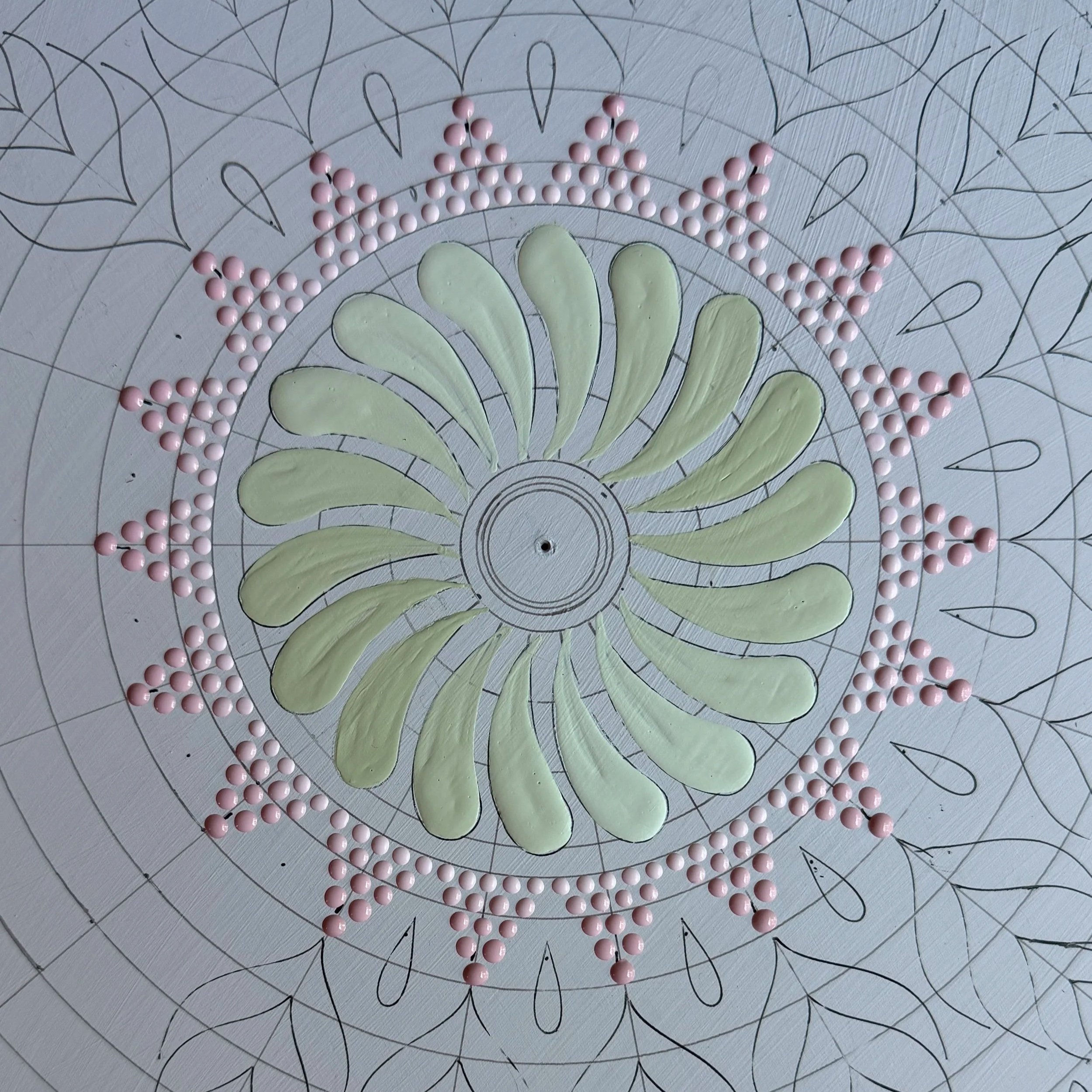 A decorative art piece featuring pink beads arranged to form a rainbow shape on a circular, hand-drawn mandala pattern with floral and leaf motifs.