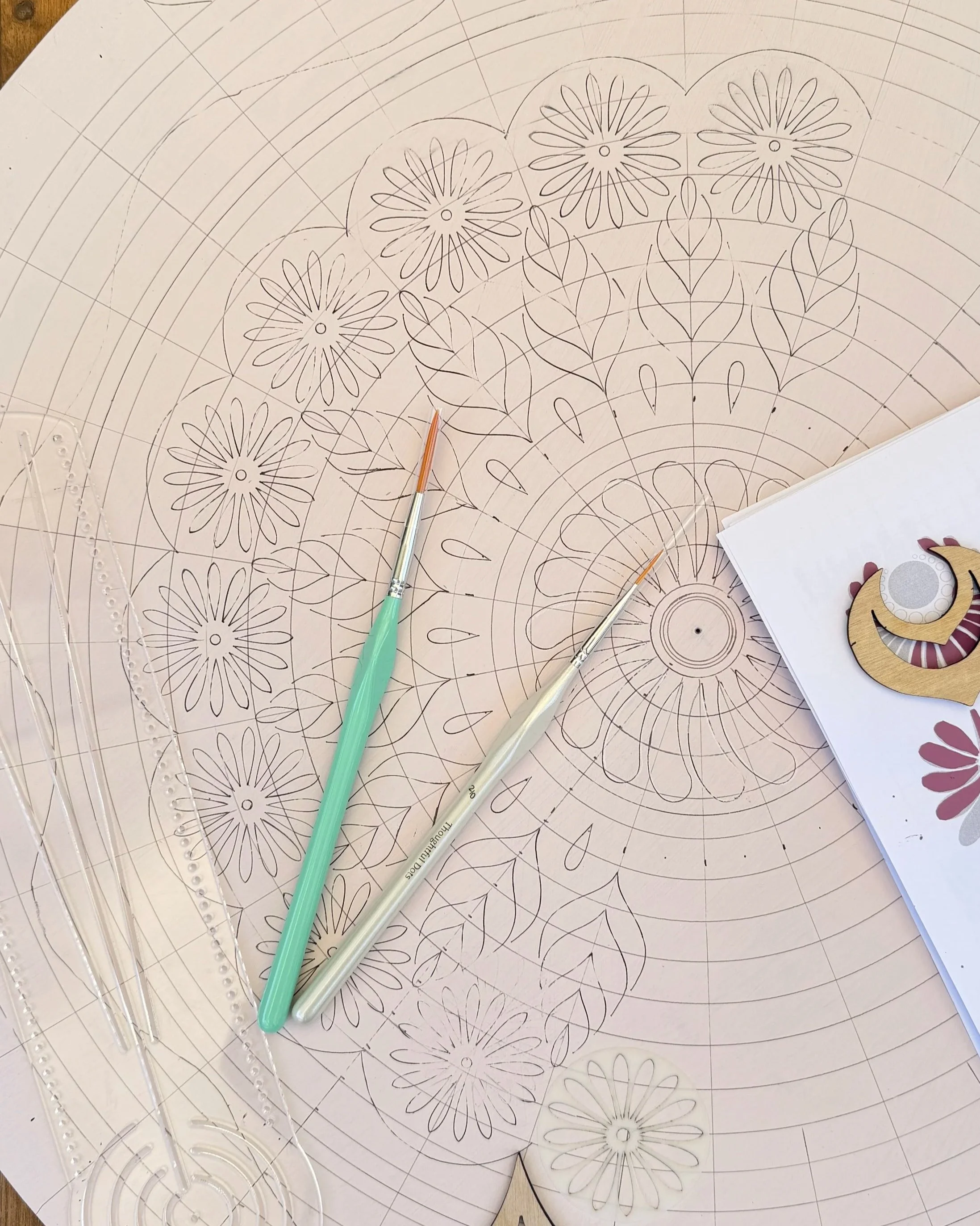A detailed design drawing with flower and leaf patterns, including circular motifs, on large sheets of paper, with two paintbrushes, a circular stencil, and a piece of finished artwork nearby.