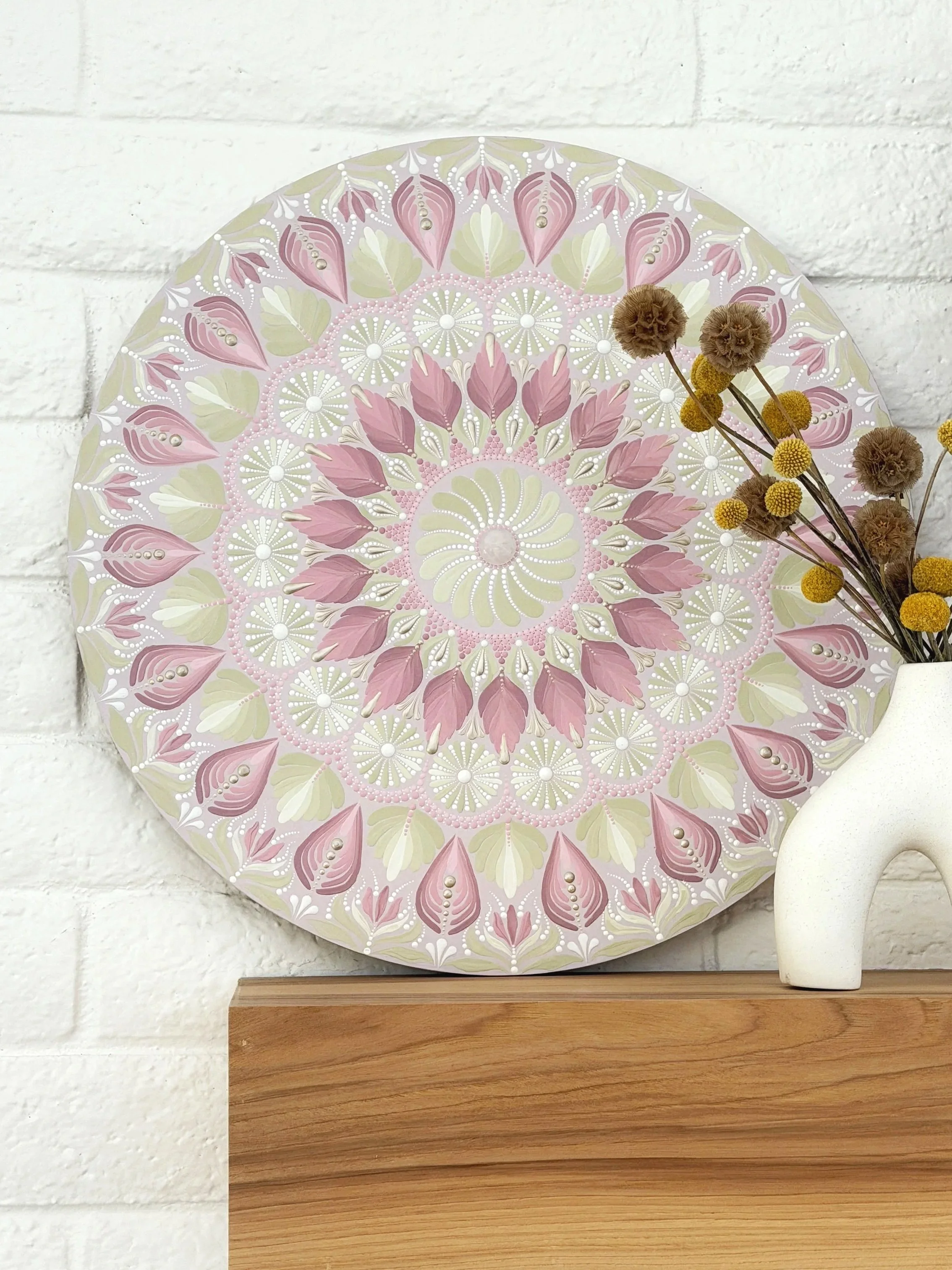 Decorative round wall art with pink and white floral pattern, a small bunch of dried flowers, and a white ceramic 'C' sculpture, against a white brick wall background.