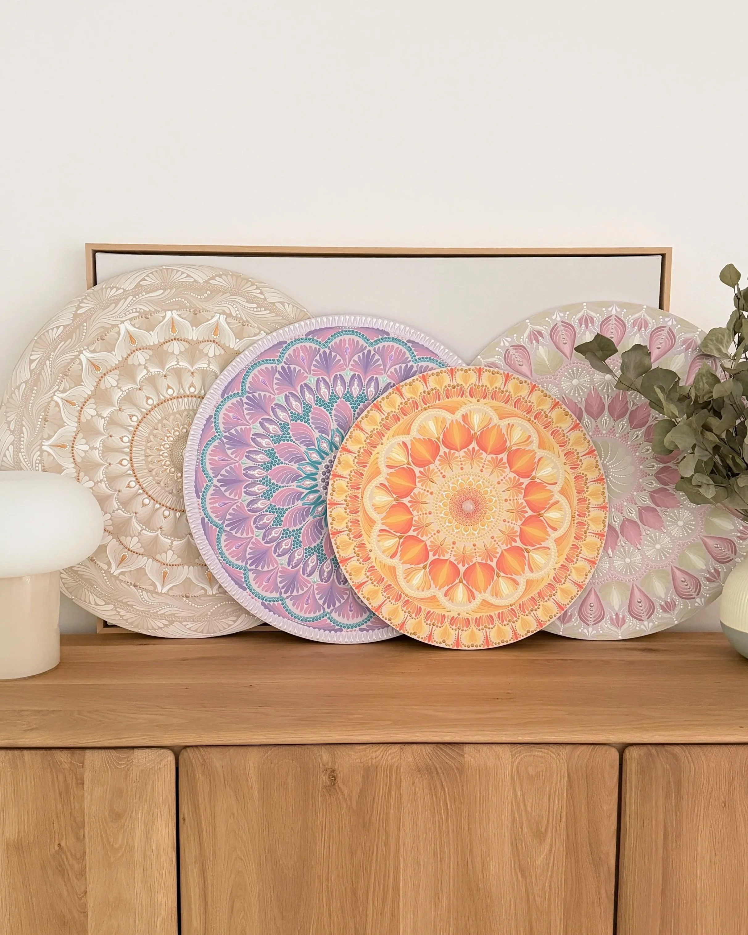 A wooden sideboard with five large decorative circular paper fans with intricate colorful floral patterns, a white vase on the left, a green leafy branch on the right, and a white wall in the background.