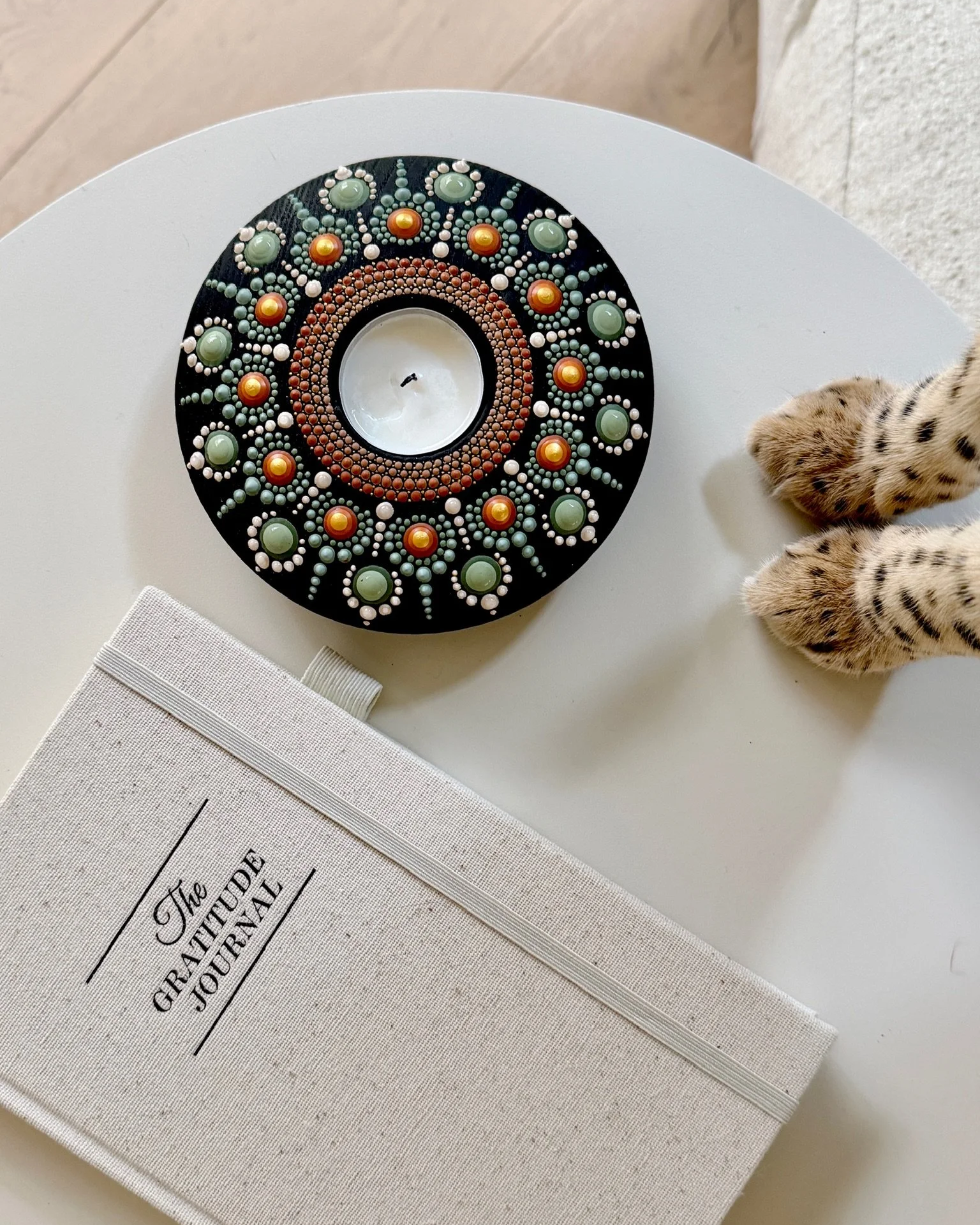 A round black decorated candle holder with a white candle inside, placed on a white table next to a closed book titled 'The Greta Guide Journal.' Part of a leopard-print cat paw is visible on the right.
