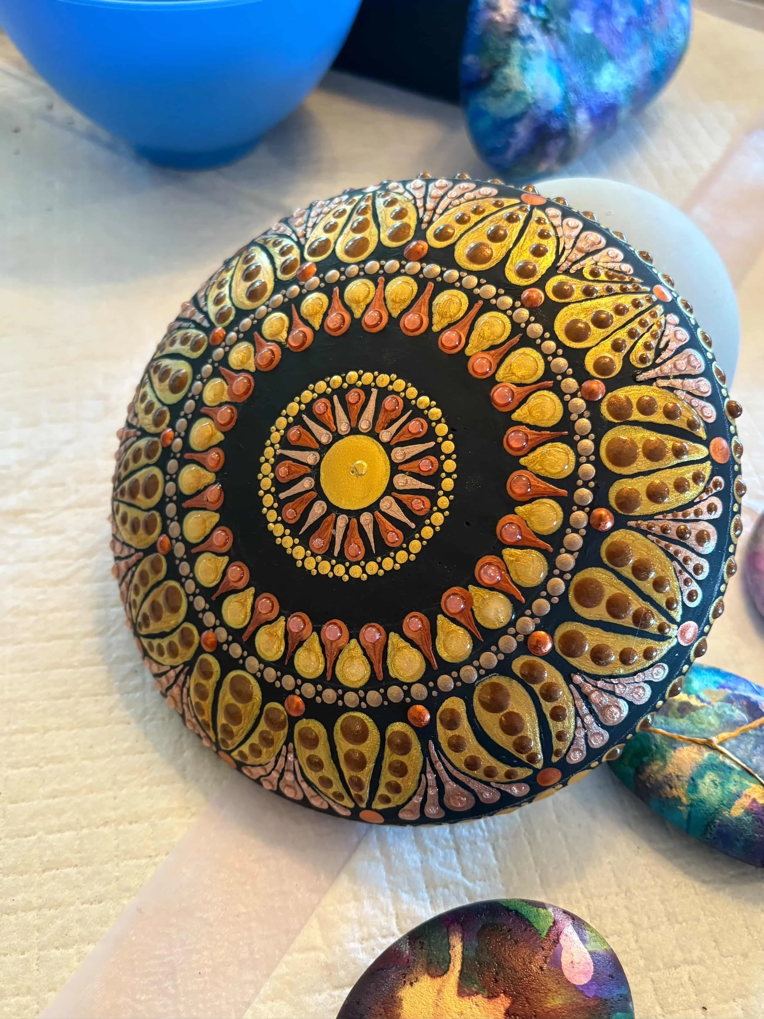 Decorative painted rock with intricate mandala design in black, gold, and orange colors.