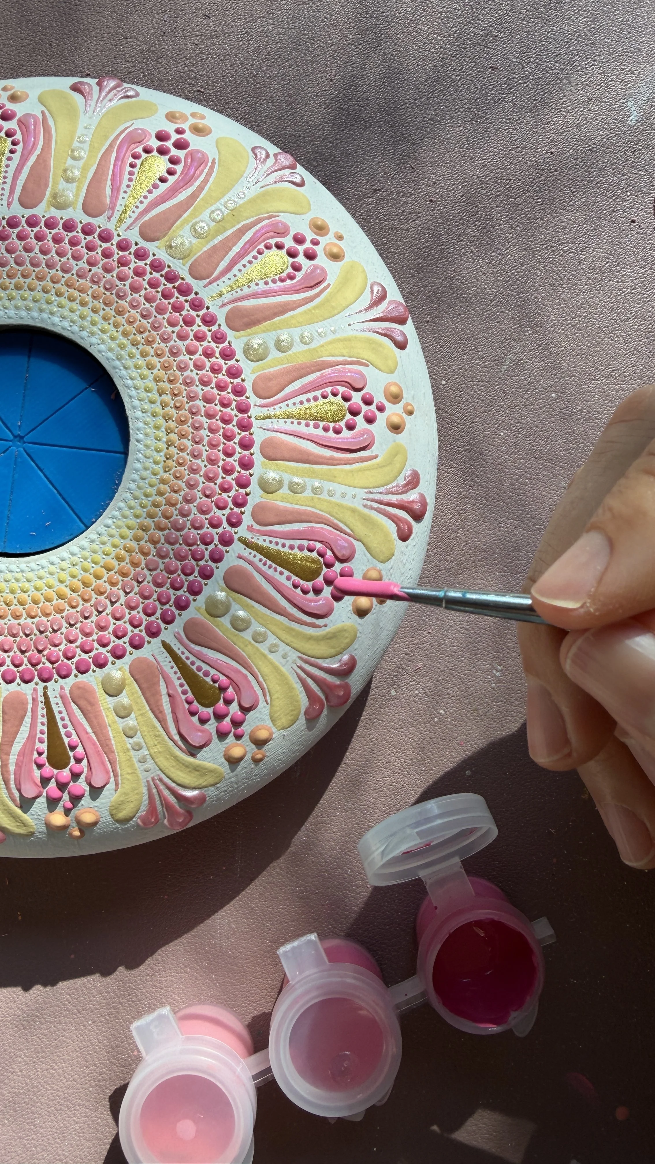A person painting a decorative, circular object with pink, yellow, and gold paint using a small brush. Several open bottles of pink paint are visible nearby.