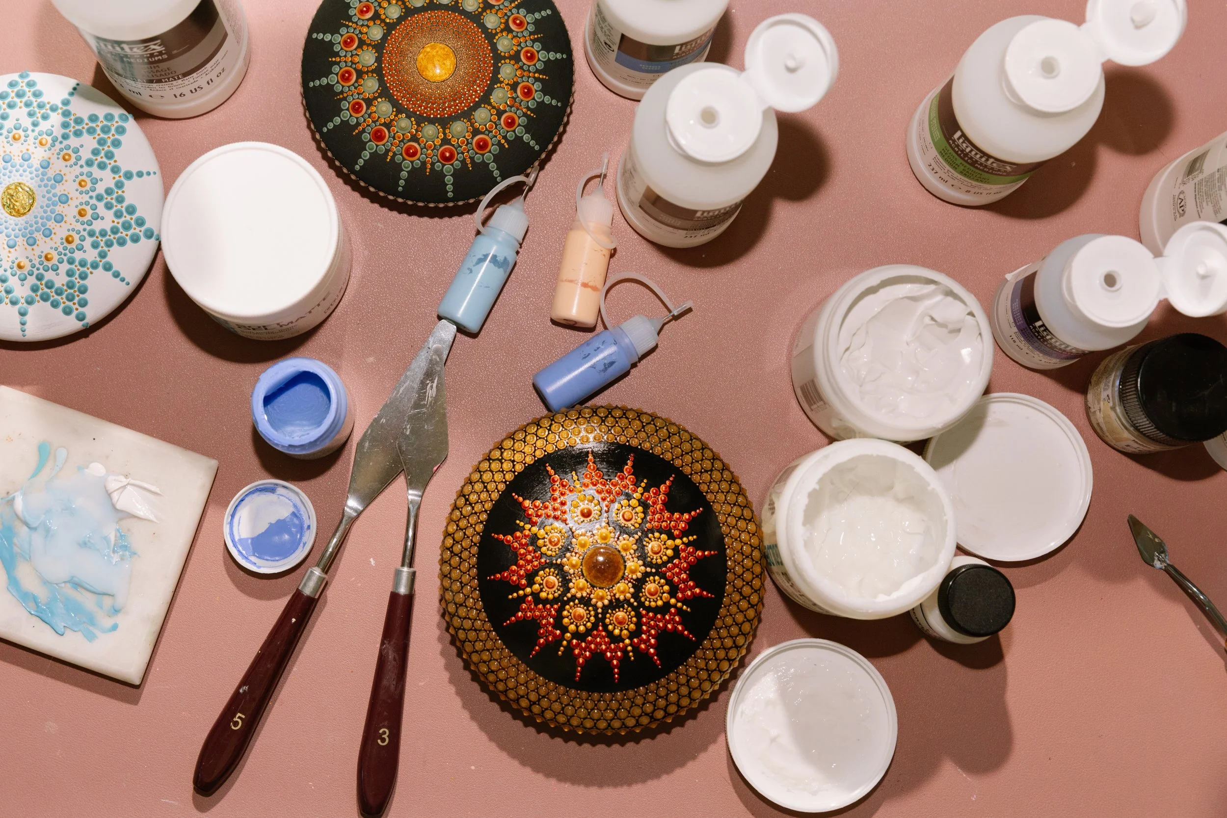 Painted stones with intricate dotwork designs, watercolor paints, brushes, and open containers of white and blue paint arranged on a pink surface, likely in the process of creating dot art on the stones.
