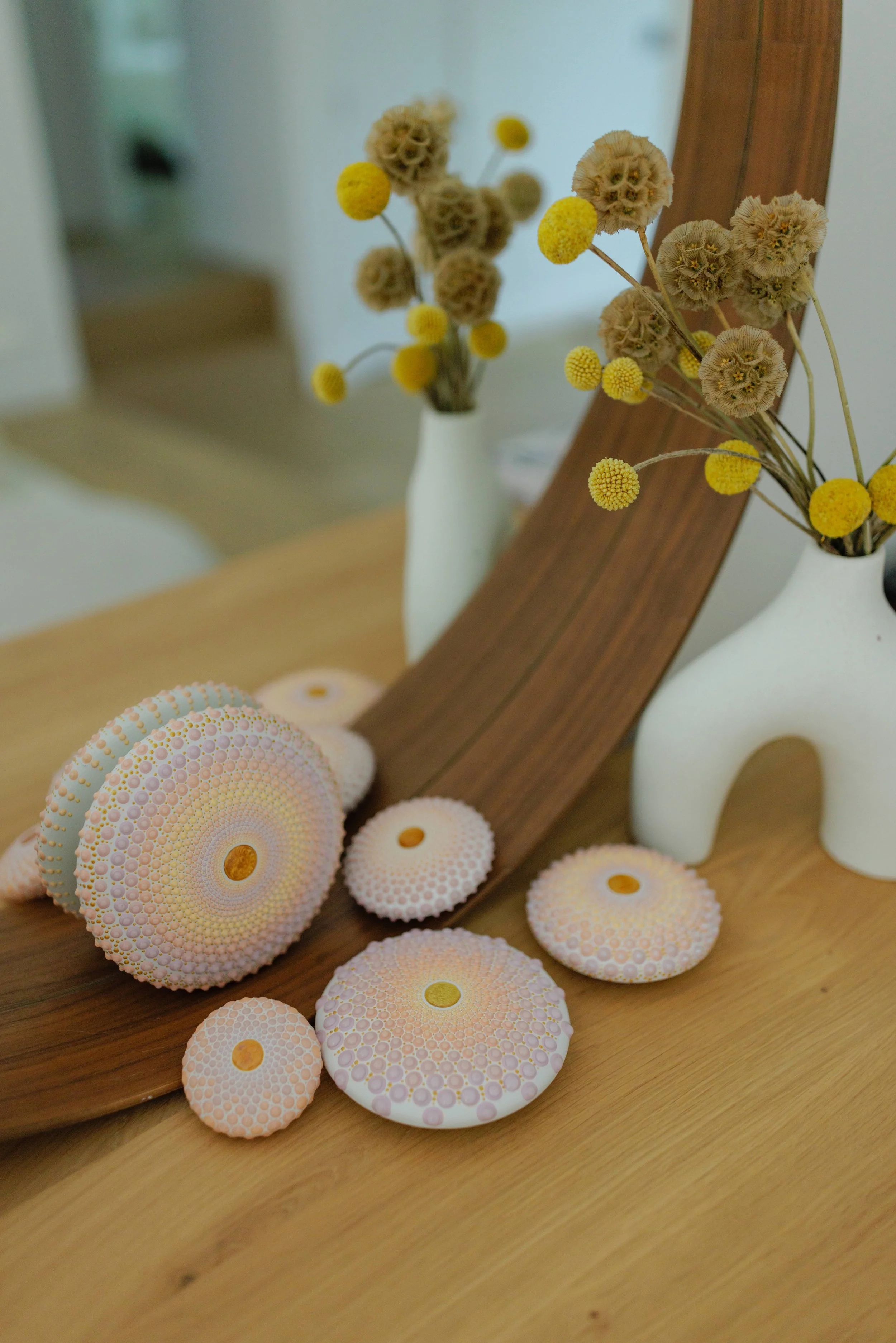 A mirror behind a vase of dried flowers and a wooden frame, with decorative circular objects on a wooden surface.