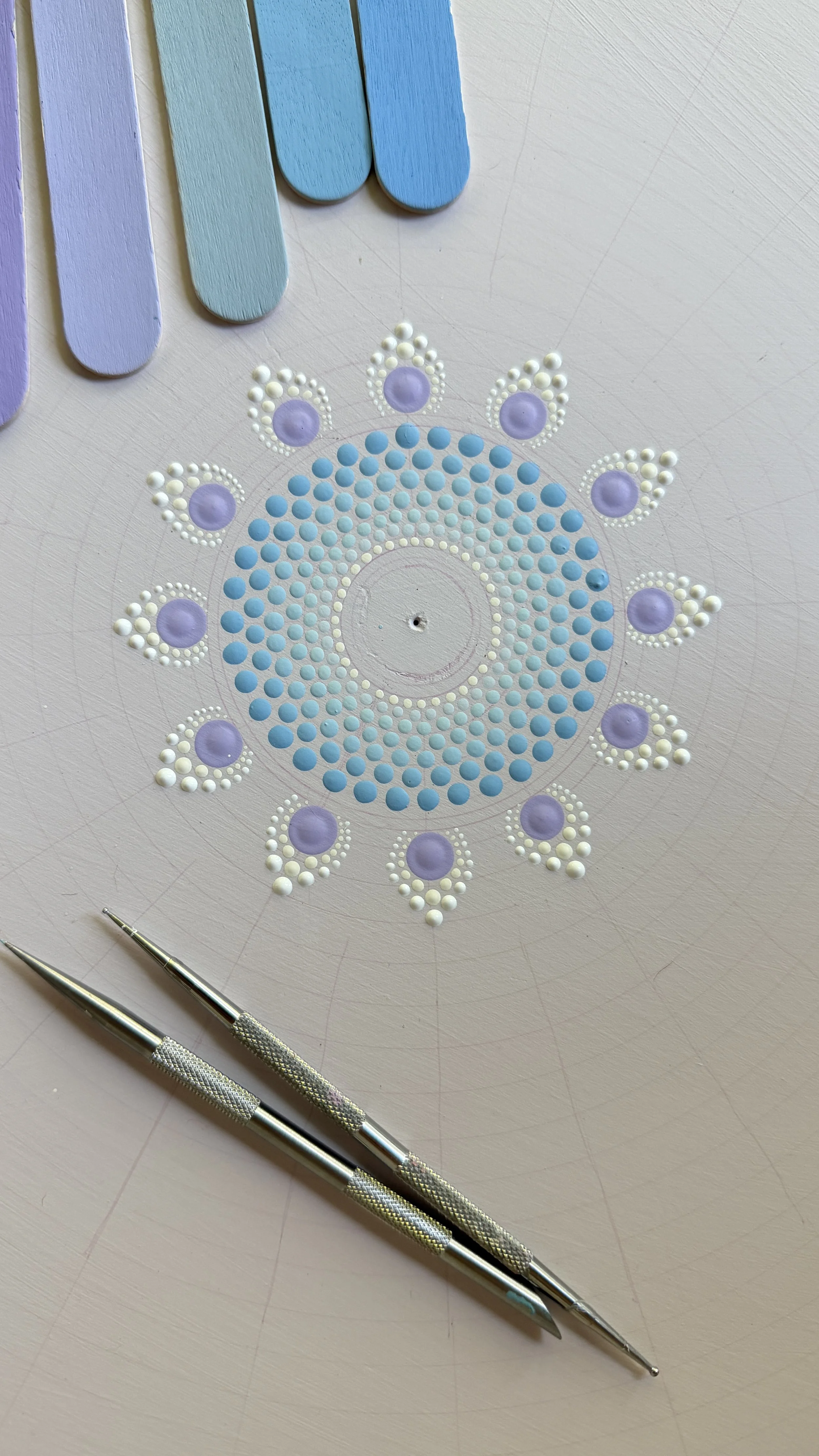A mandala design created with pastel-colored dots and shapes on a work surface, accompanied by two metal dotting tools in the foreground.