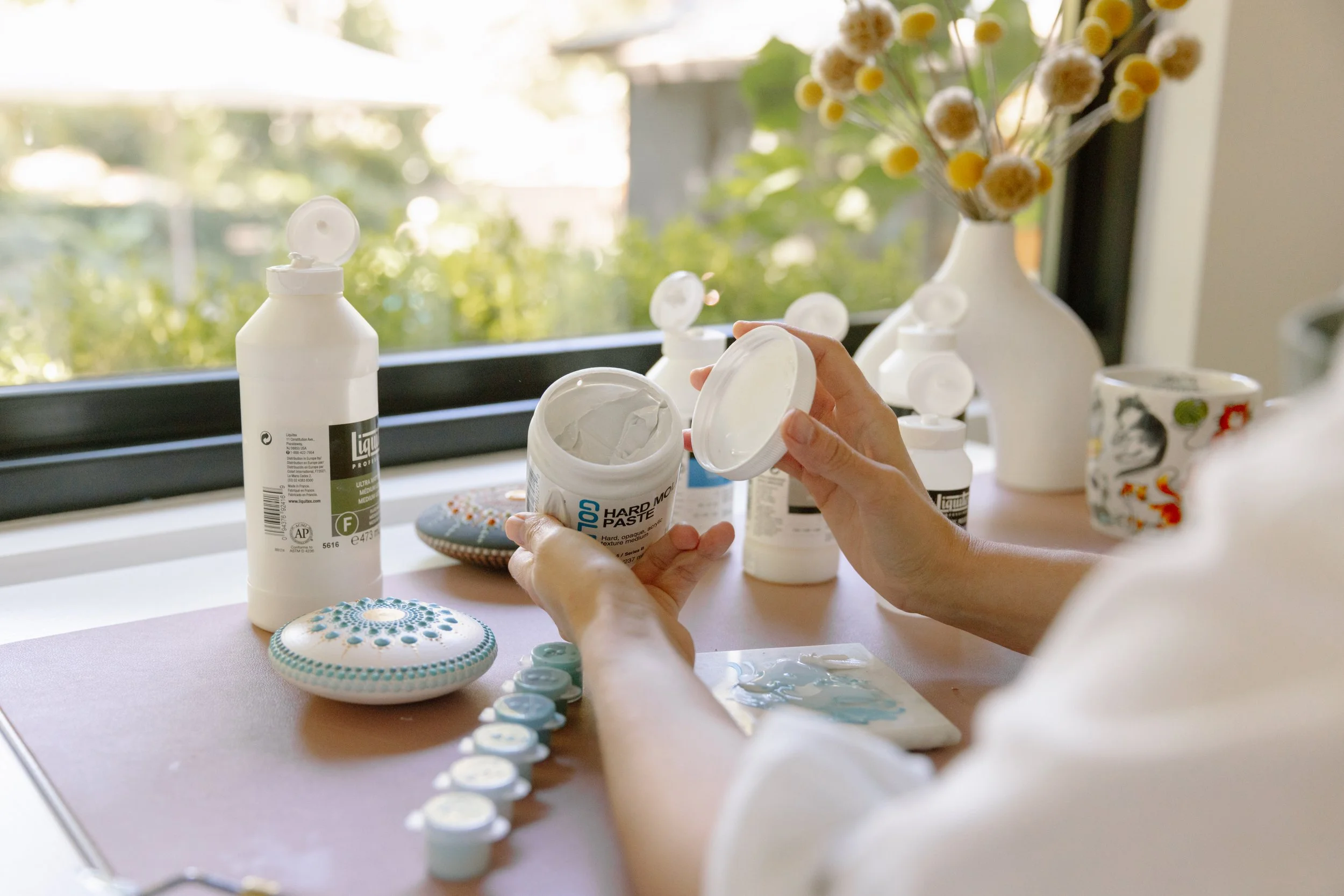 Someone opening a container of white modeling clay or similar material on a wooden surface, with decorative buttons and painted stones nearby. In the background, there are bottles of paint and a vase with yellow and white flowers, with sunlight comin
