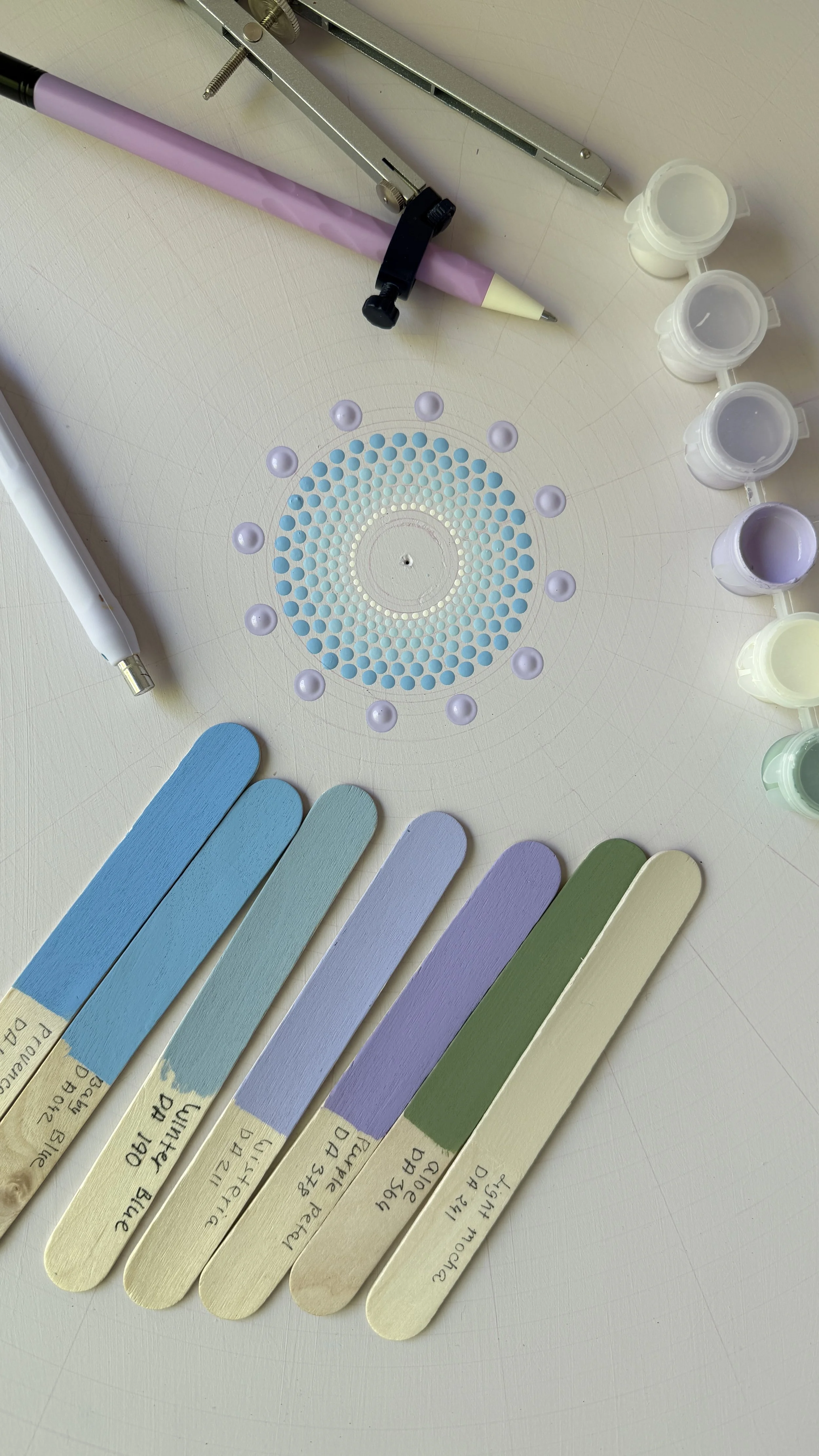 Color swatches labeled with paint names, ranging from blue to green, a circle of lavender paint dollops, a purple mechanical pencil, a metal compass, and small paint containers.