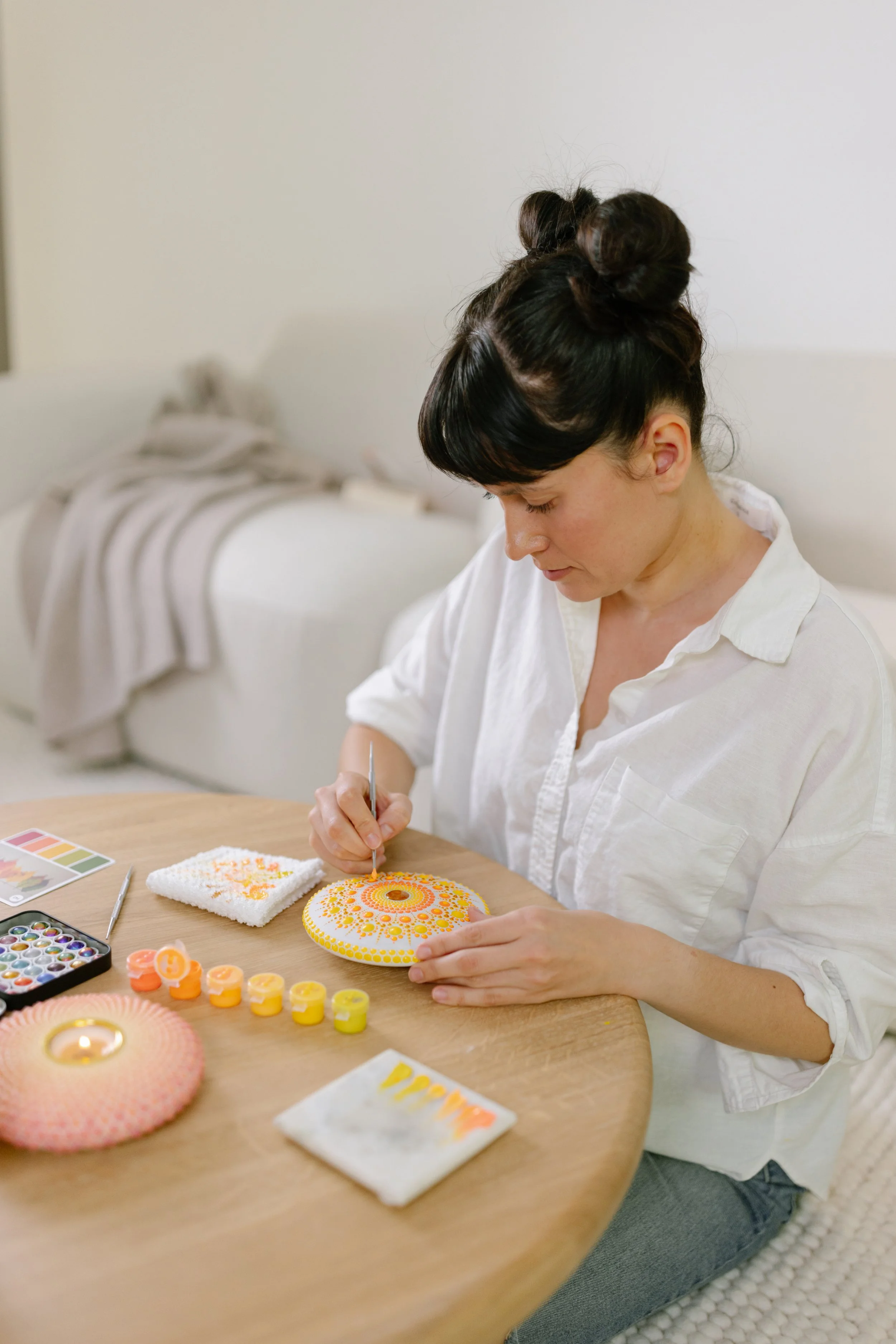Woman sitting at a wooden table creating dot mandala art using paints and tools. The table has watercolor paints, a painted mandala object, and a color palette. She is wearing a white blouse, and the background shows a white wall and a bed with blank