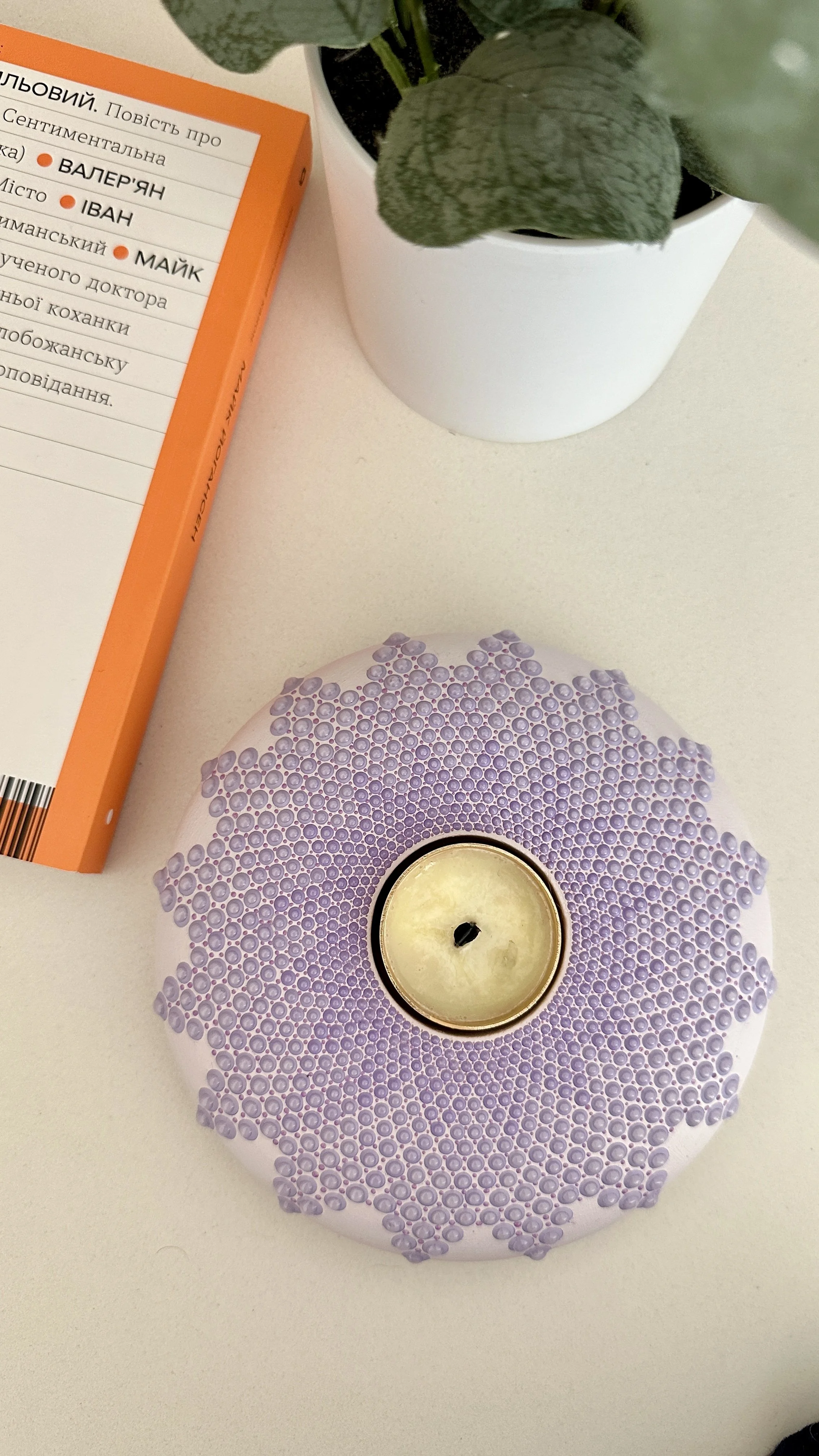 A white circular candle holder decorated with purple beads surrounding a yellow candle, a potted plant with green leaves in a white pot, a book with orange and white cover with text, and part of a purple-patterned object on a white surface.