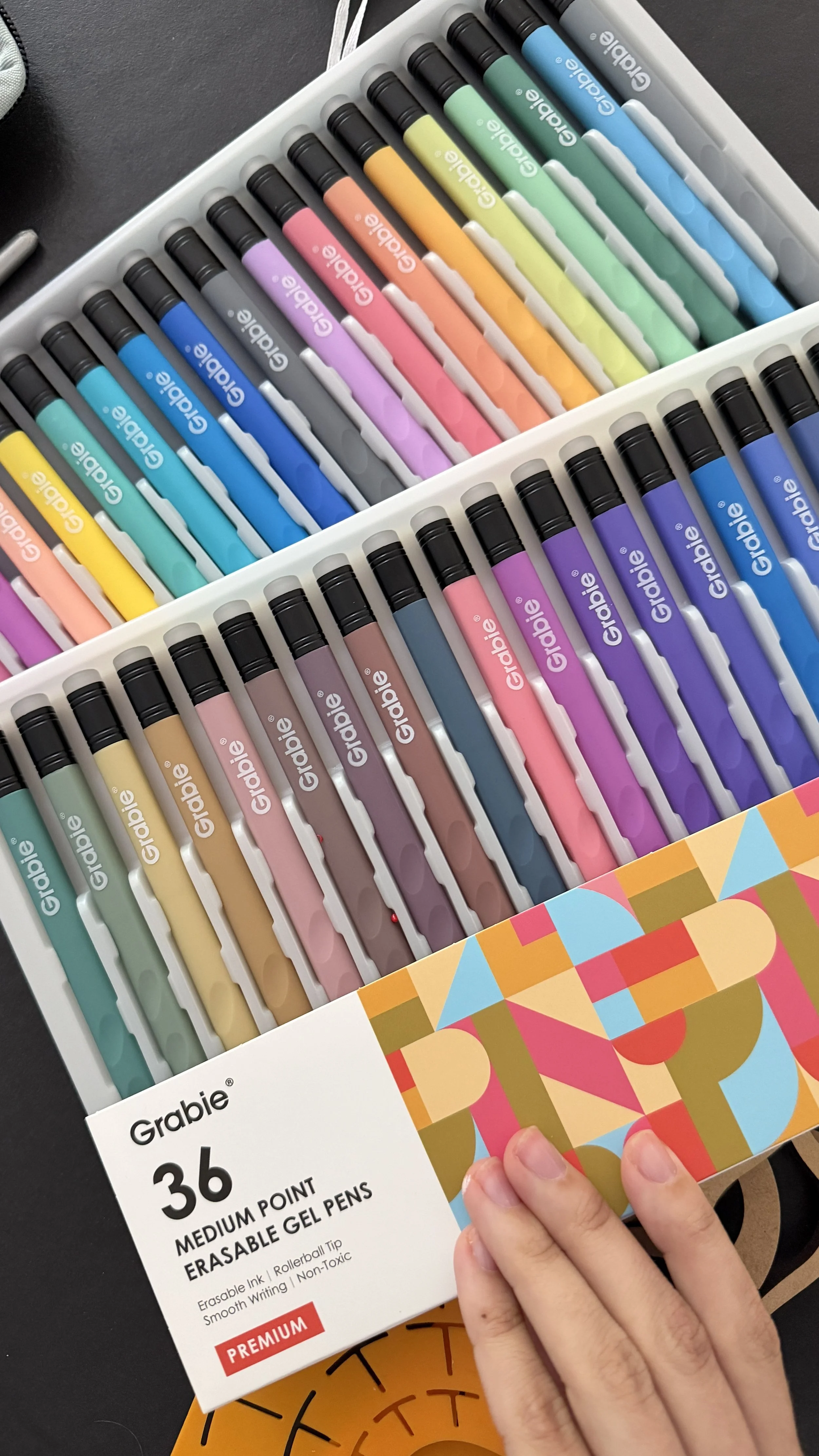 A box of 36 Grable medium point erasable gel pens in various pastel colors, held by a person's hand with a colorful geometric patterned sleeve. The pens are arranged neatly in a plastic tray inside the box.