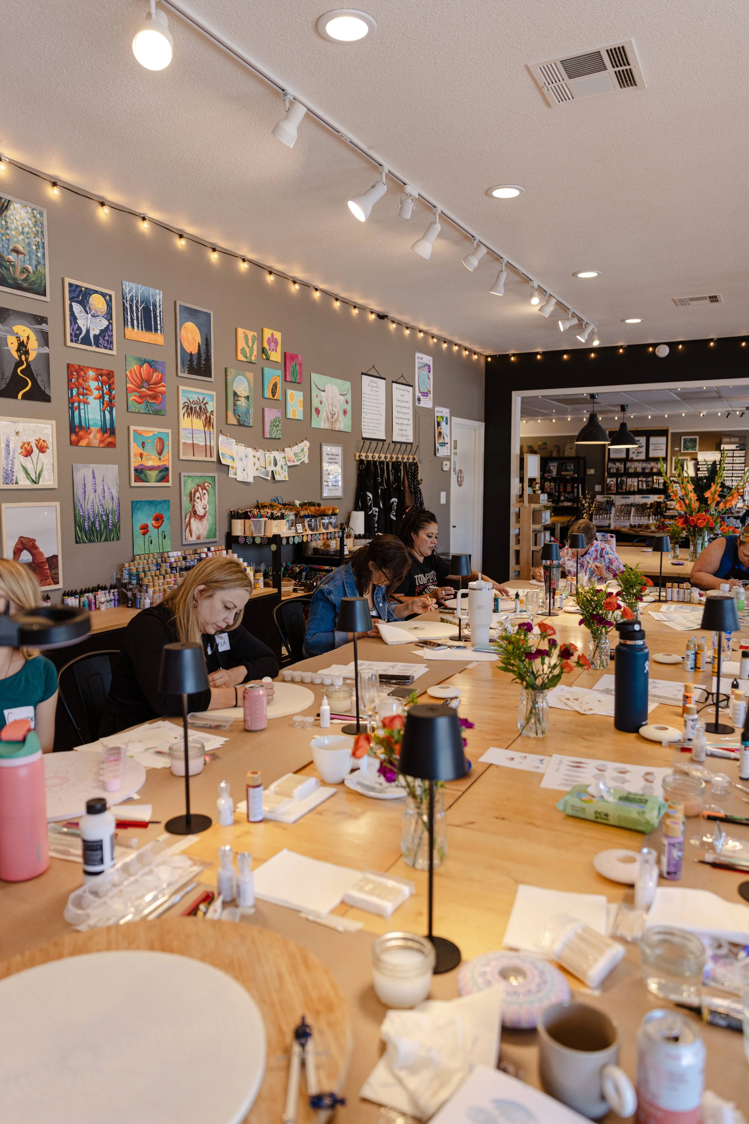A group of people sitting at a large wooden table involved in arts and crafts, with various art supplies, flowers, and artwork displayed on the wall in a well-lit room.