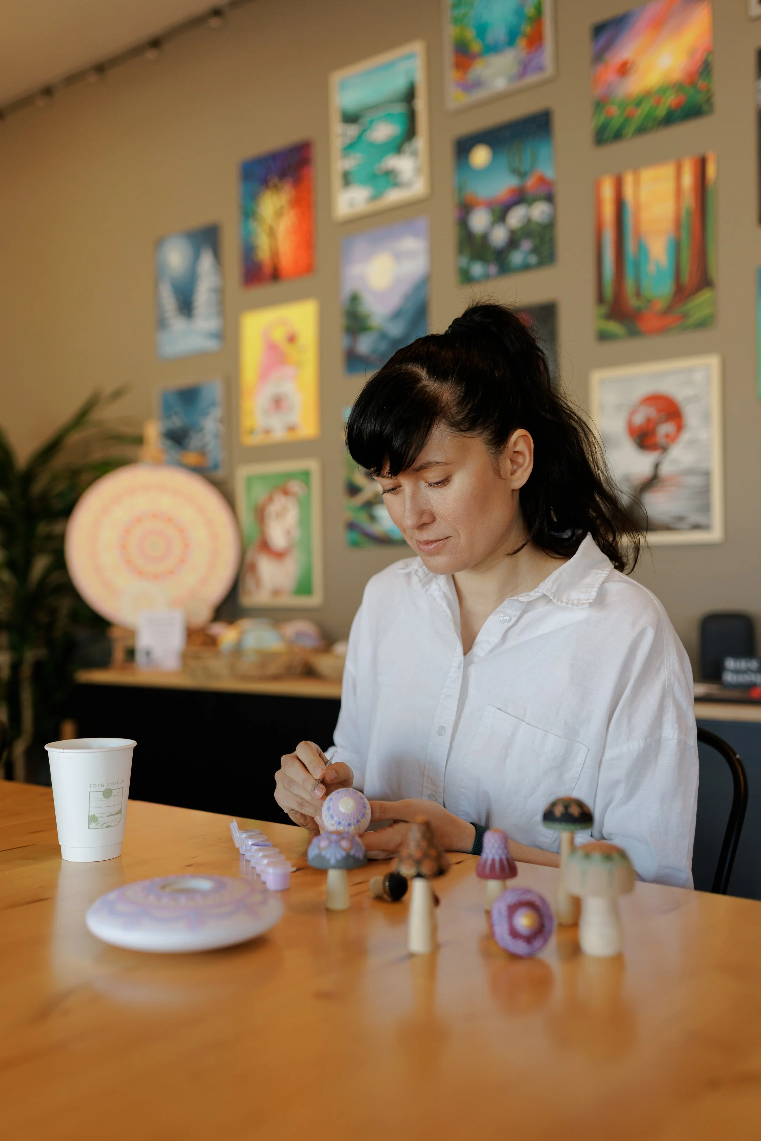 A woman with black hair and a white shirt decorated with various small colorful objects is sitting at a wooden table in an art gallery or studio. Behind her on the wall are various colorful paintings, including landscapes and abstract art. The table 