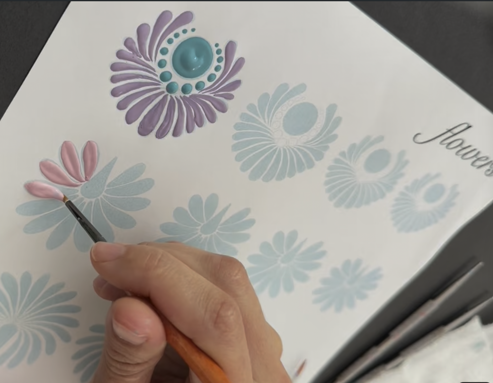 A person is painting flower illustrations on paper, using pink and purple colors, with a paintbrush in hand. The paper has flowers with text that reads 'Flowers'.