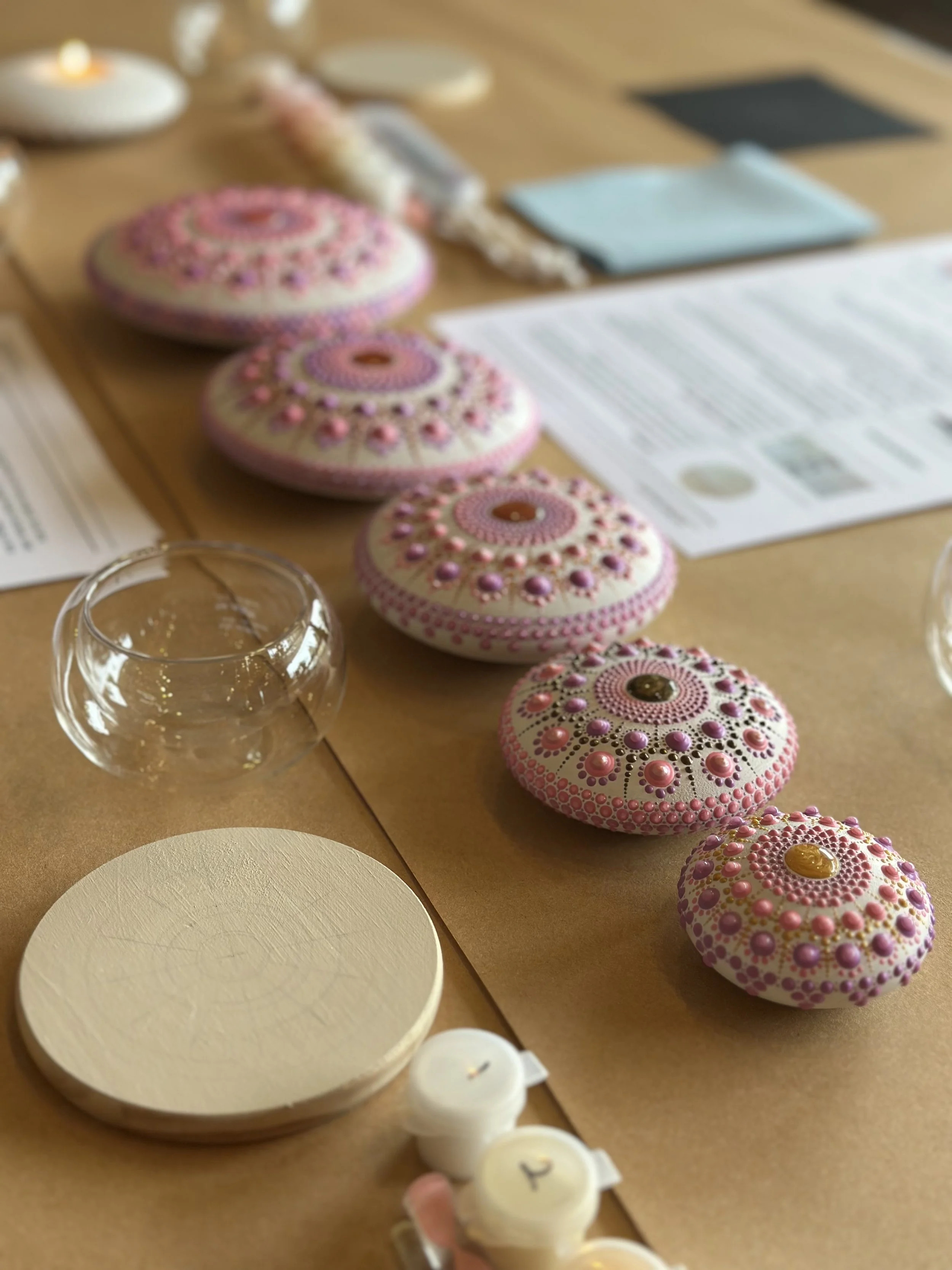 Decorated pink and white rocks with patterns on a table, along with clear glass cups, white candles, and papers.