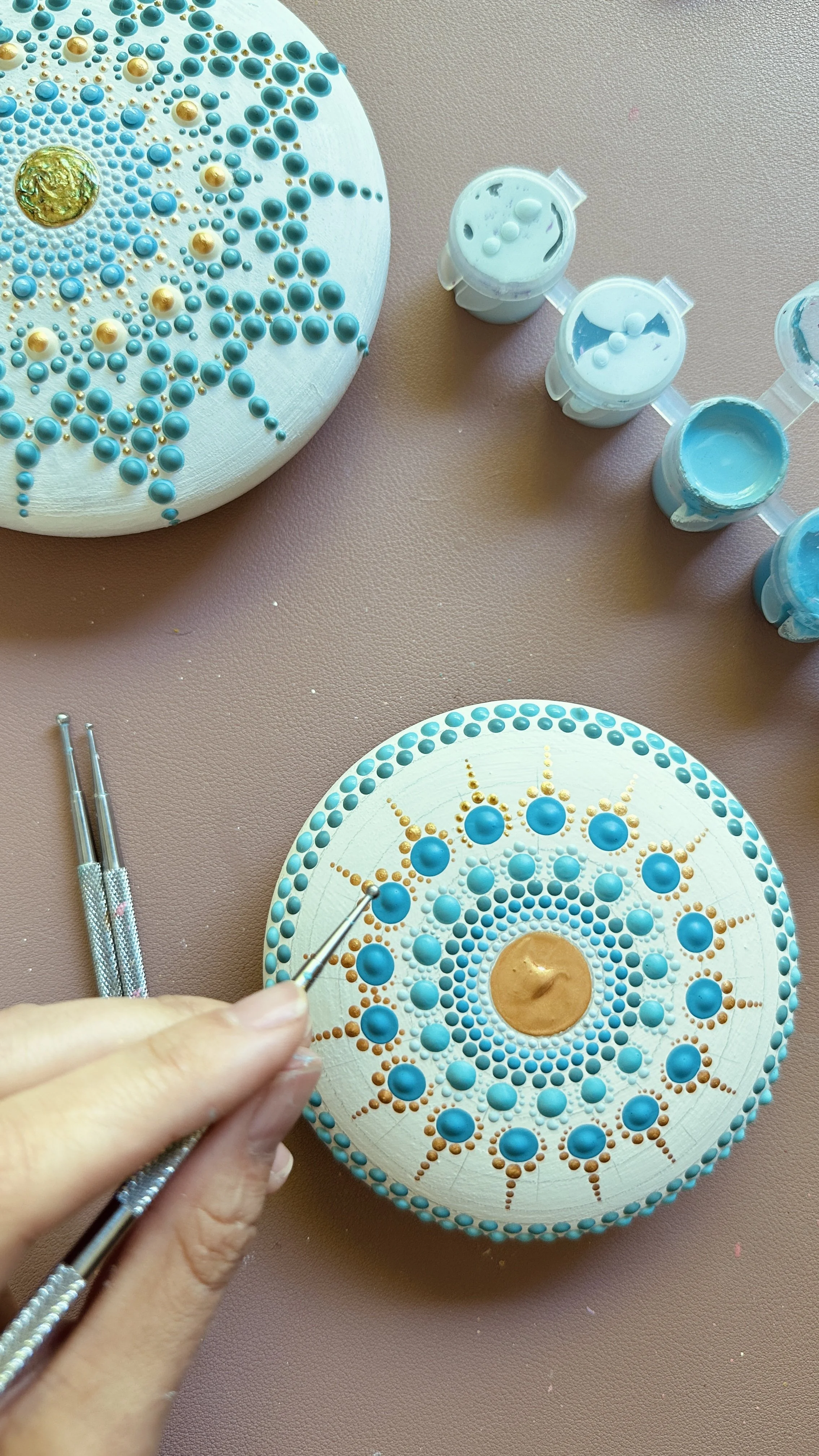 Close-up of a person decorating a circular wooden art piece with blue and gold paint dots, using dotting tools, with small containers of blue paint nearby.