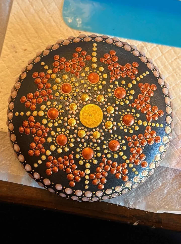 Painted rock with a mandala design made of dots in red, yellow, and pink on a black background.