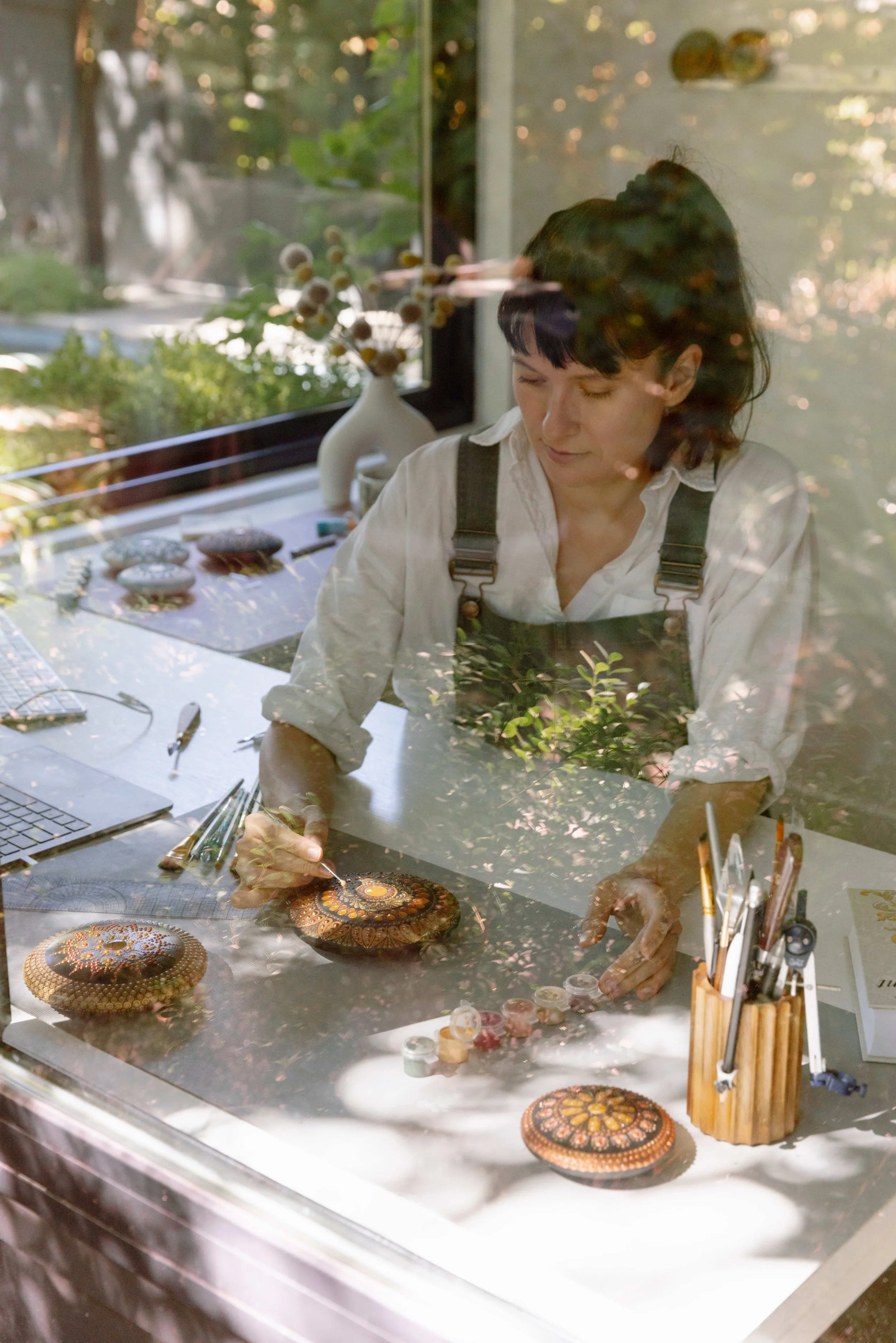 An artist working on circular dot paintings through a glass window, with outdoor scenery and reflections visible.