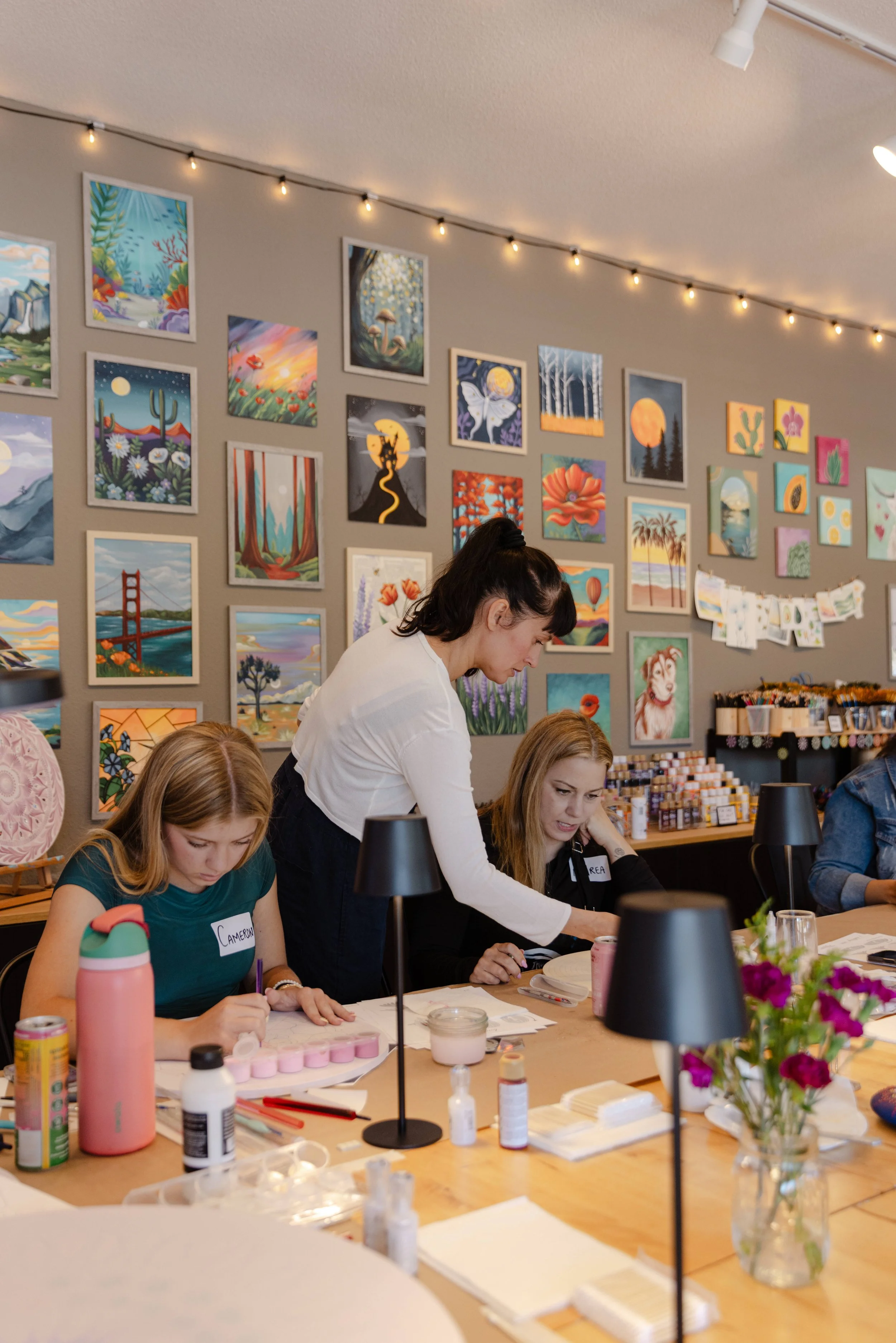 People participating in an arts and crafts activity at a table with various supplies, in a room decorated with colorful landscape and nature paintings on the wall, and string lights overhead.