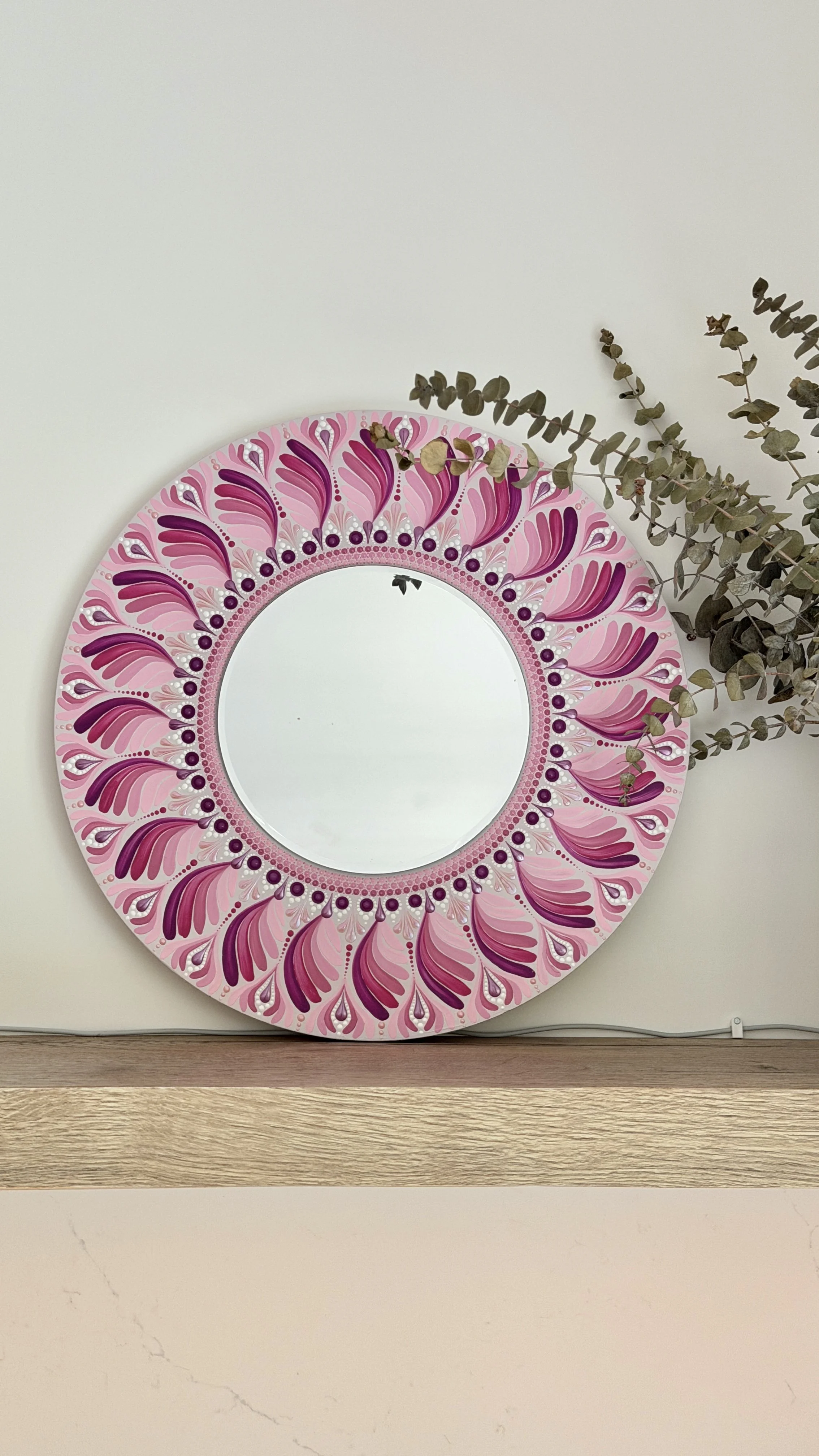 Decorative round mirror with a pink, purple, and white floral pattern border hanging on a white wall, with a sprig of dried eucalyptus to the right of the mirror.