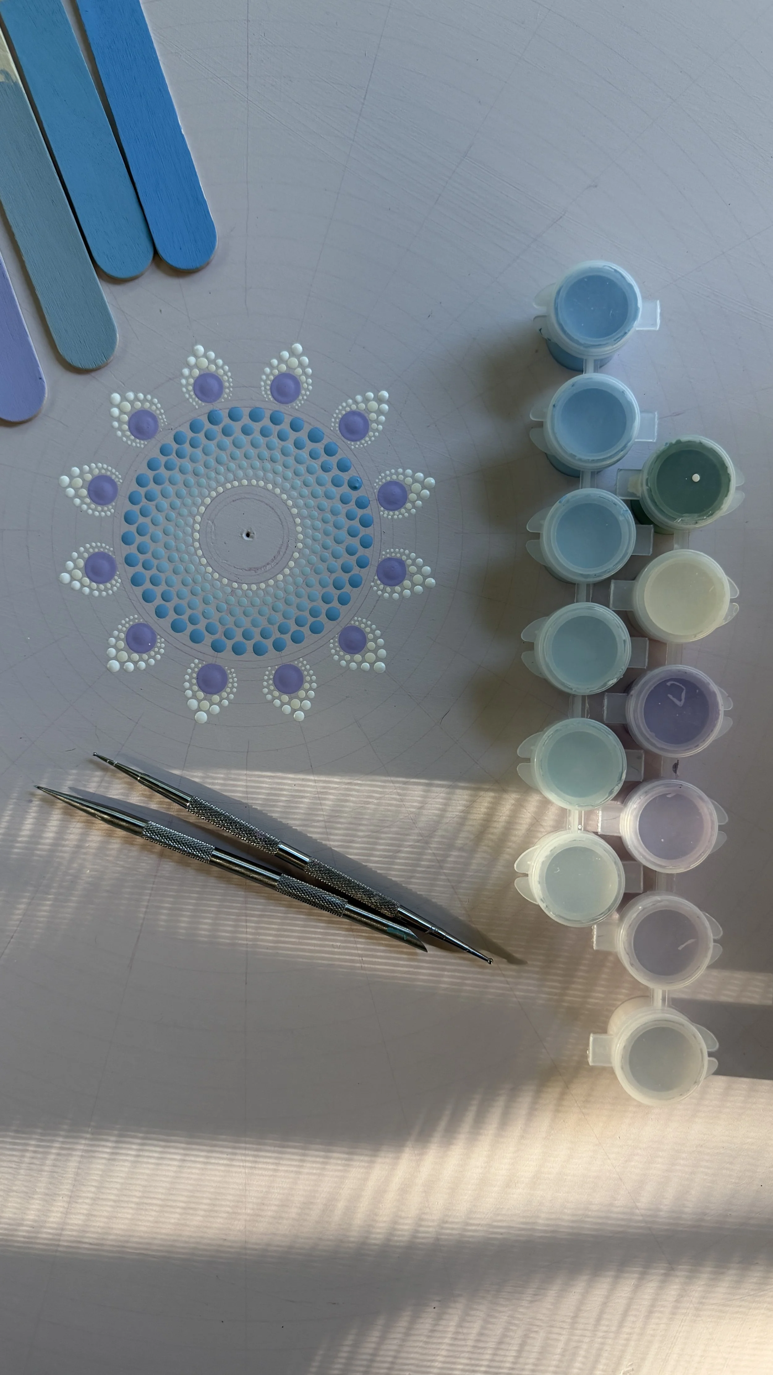 Nail art design tools on a white surface, including a dotted nail art wheel, small paint pots in various shades of blue and white, and two metal nail dotting tools.