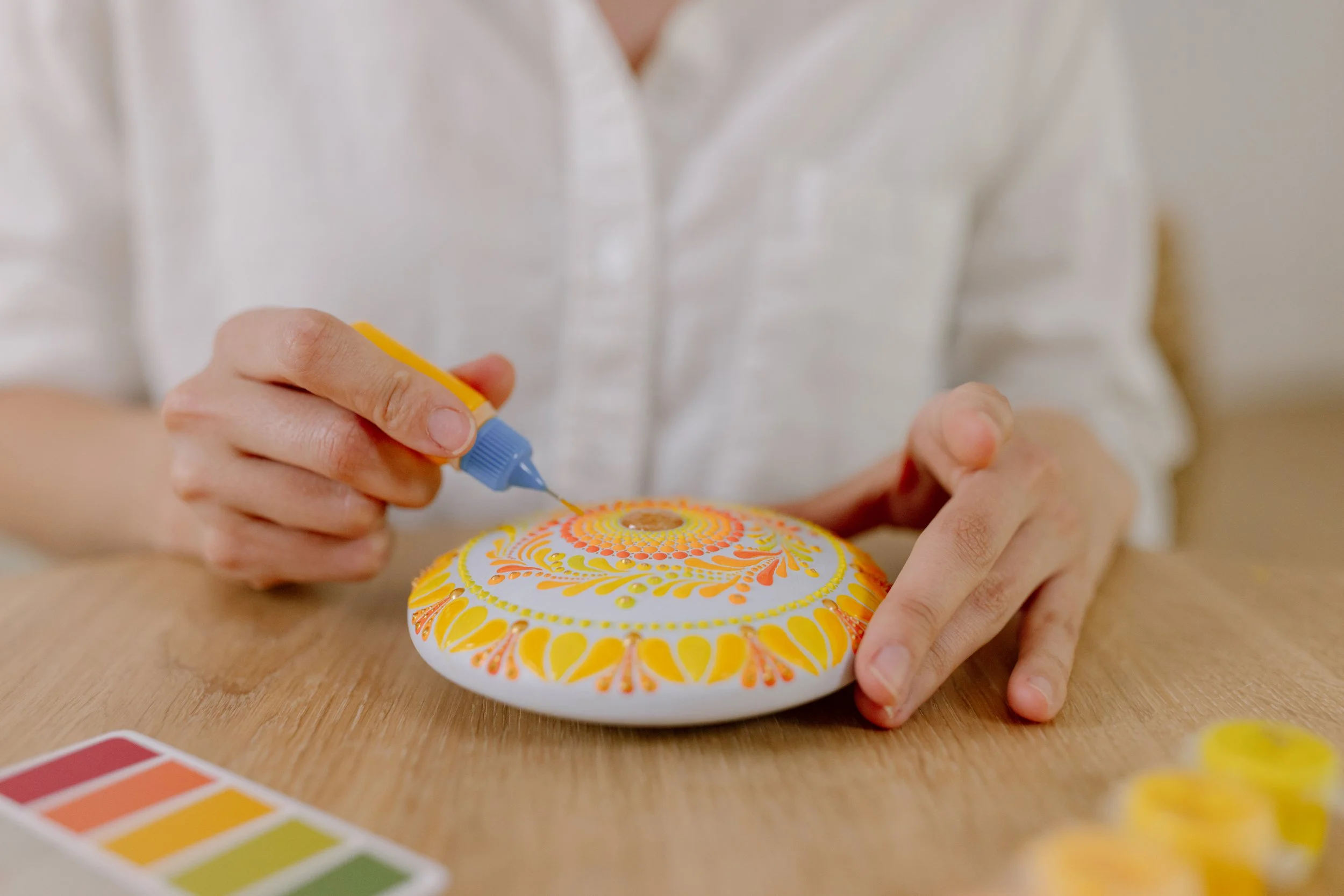 Person decorating a decorative object with yellow and orange patterns using a small tube of paint.