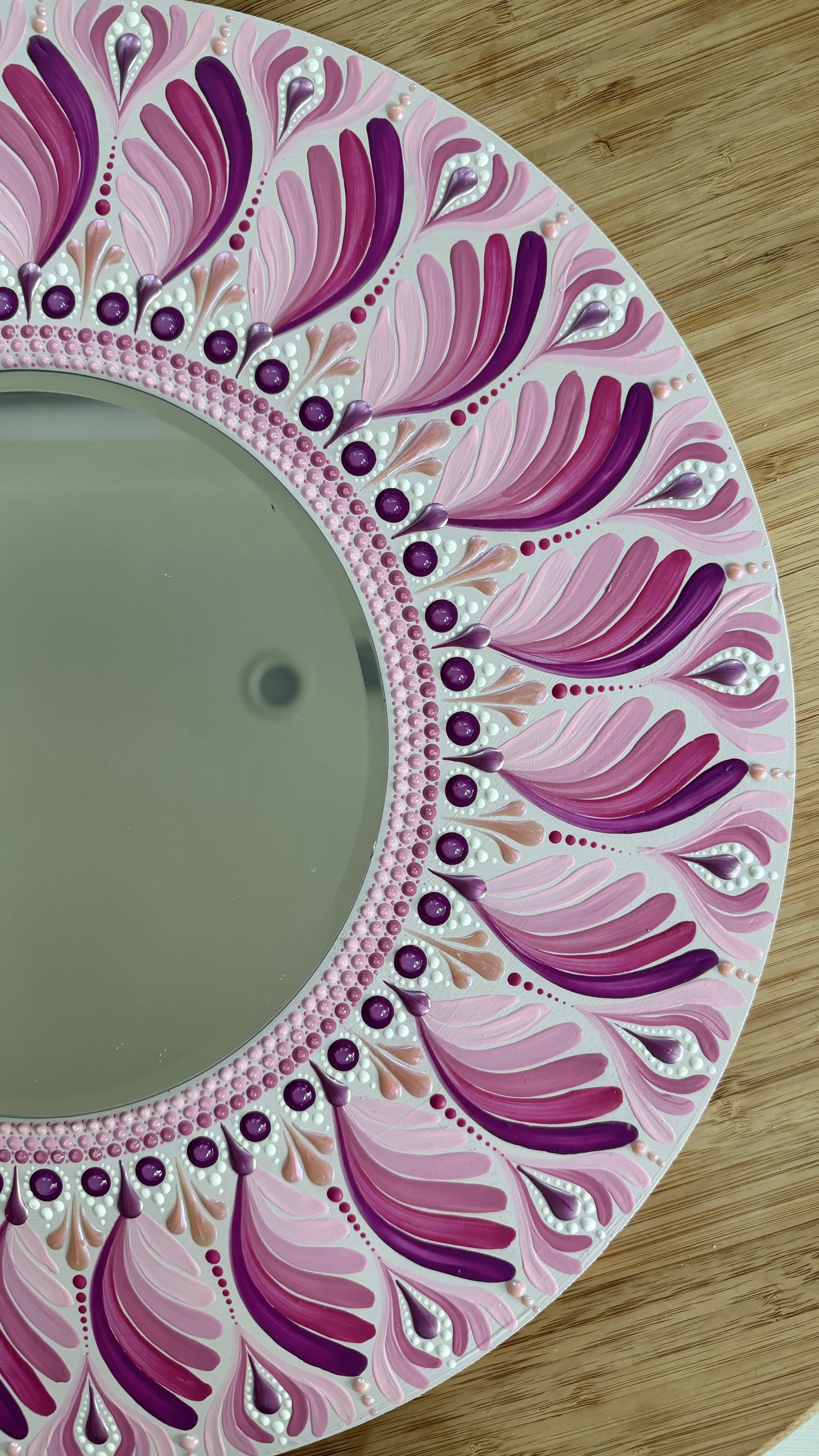 Decorative round mirror with a pink, purple, and white ornate border, placed on a wooden surface.