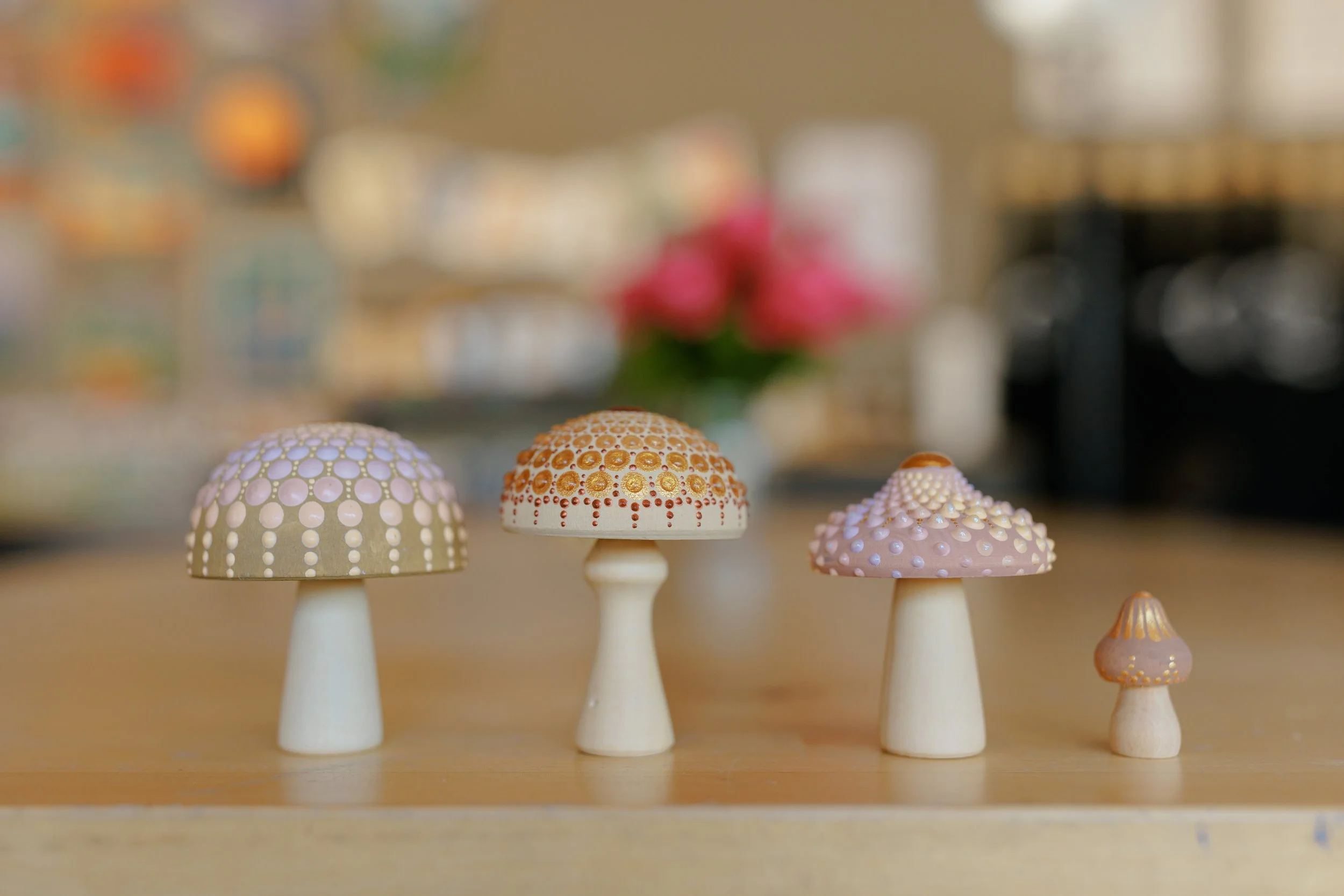 Collection of decorative mushroom-shaped objects with patterned caps in pastel colors, arranged on a wooden surface with blurred background featuring colorful objects and a plant with pink flowers.
