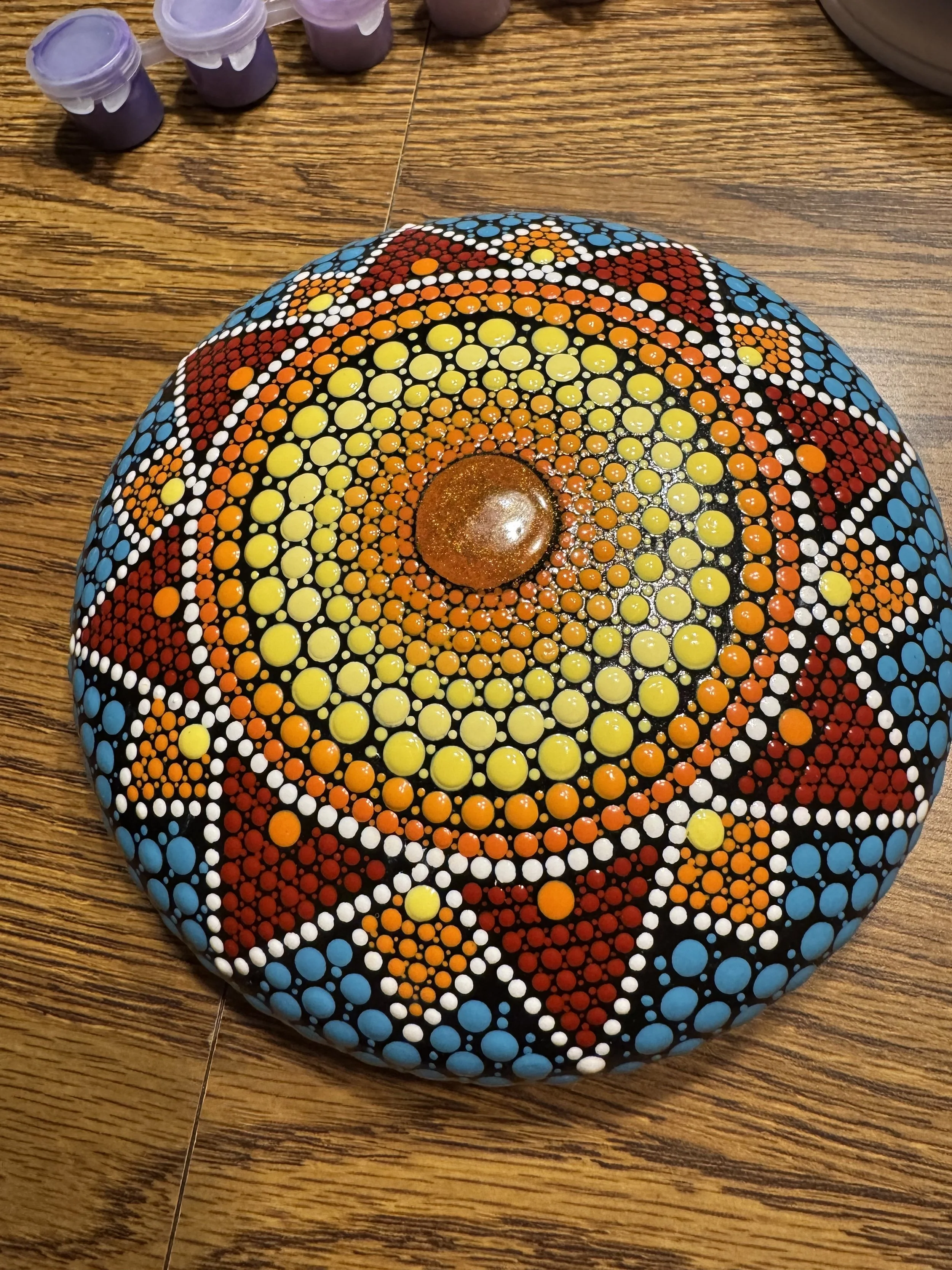 Colorfully painted rock with concentric dot patterns in yellow, orange, white, red, blue, and black, placed on a wooden surface.