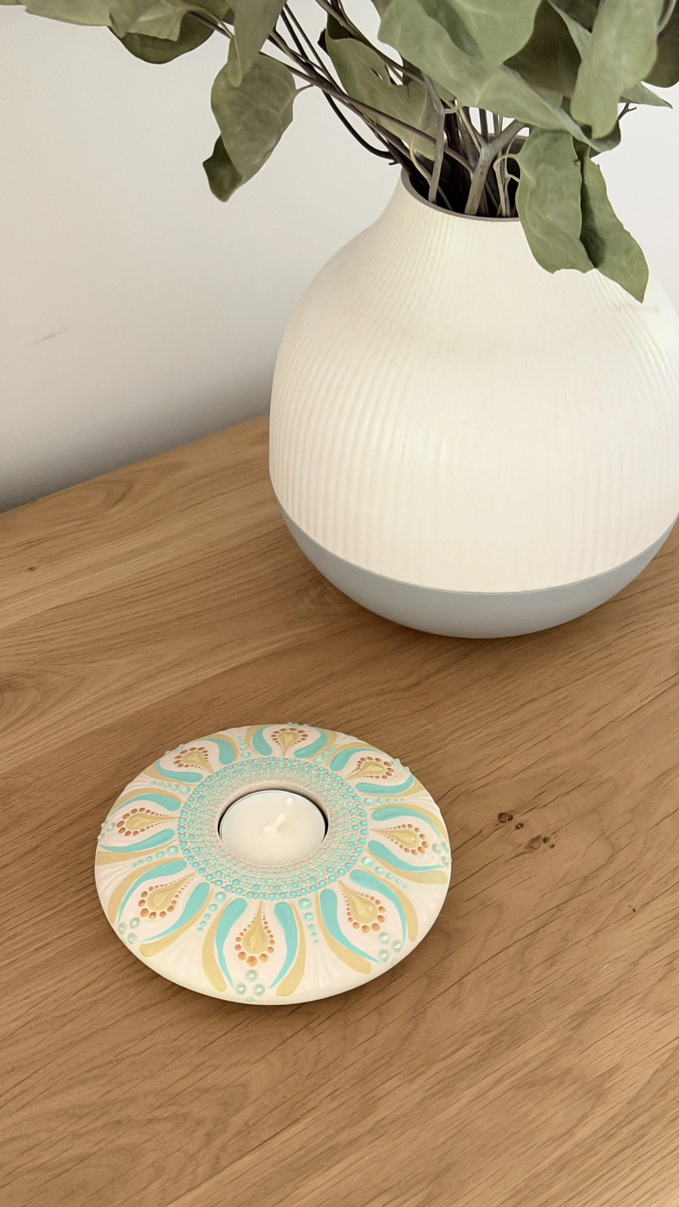 A decorative candle holder on a wooden table with a patterned design in white, blue, and gold, and a ceramic vase with green leaves above it.