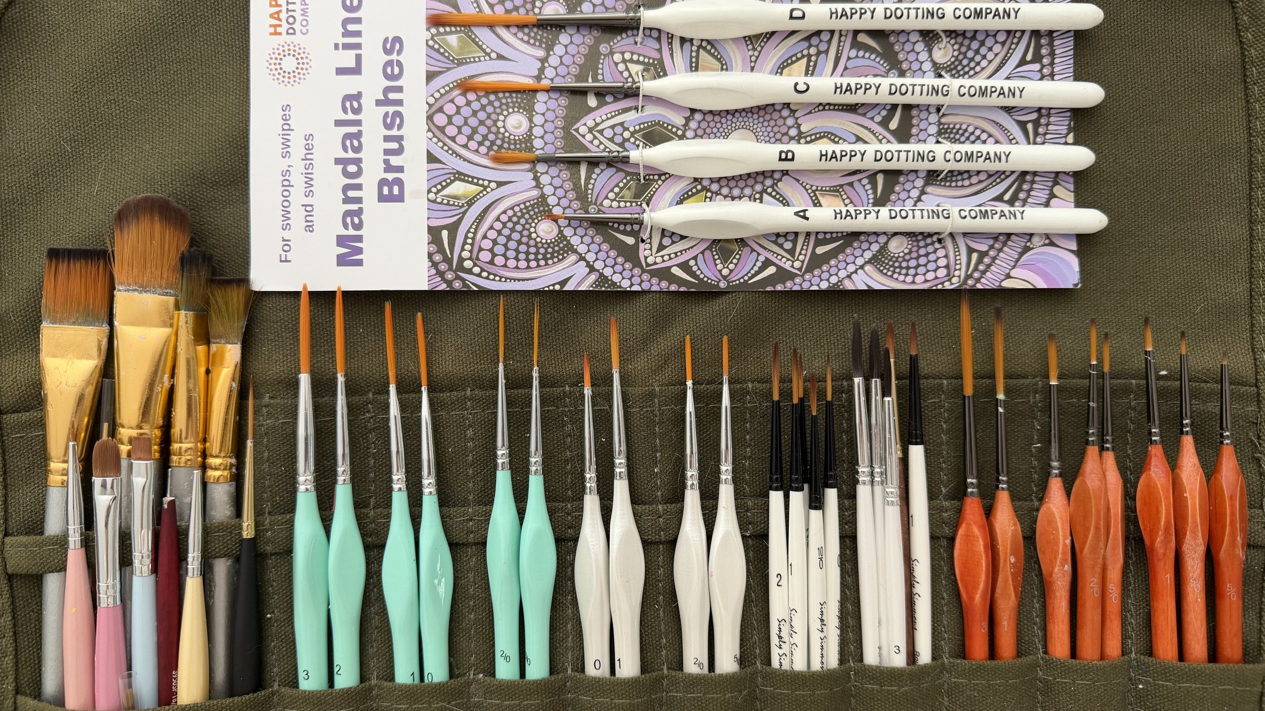 A set of multi-colored paint brushes and dotting tools arranged neatly on a fabric surface, with a decorative mandala design on a packaging label at the top.