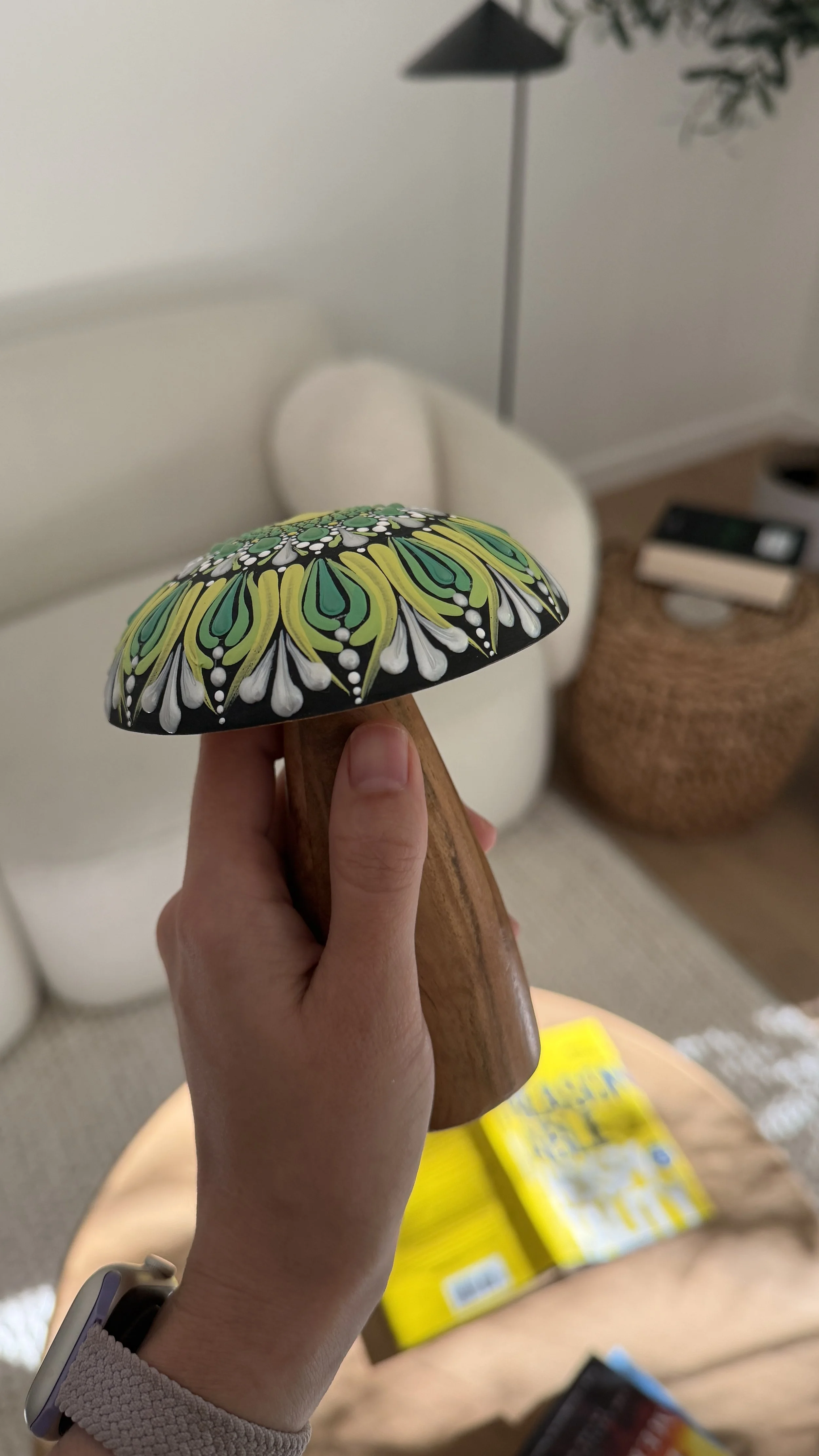 Close-up of a colorful painted wooden mushroom decoration being held by a person's hand, with a sofa, a floor lamp, a basket, and some snack boxes in the background.