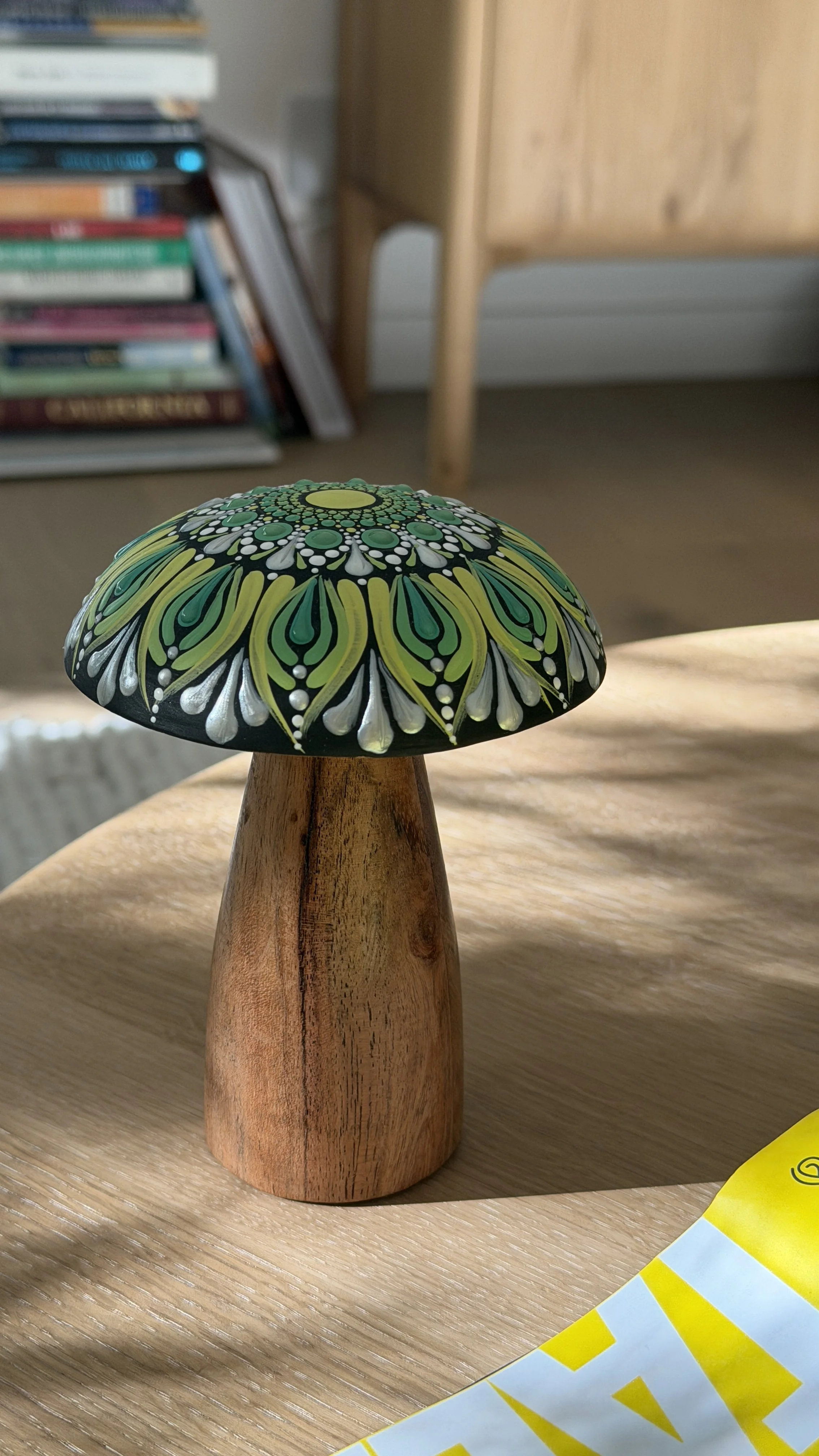 Painted decorative mushroom figurine with black, green, yellow, and white patterns on the cap, mounted on a wooden base, placed on a wooden surface.