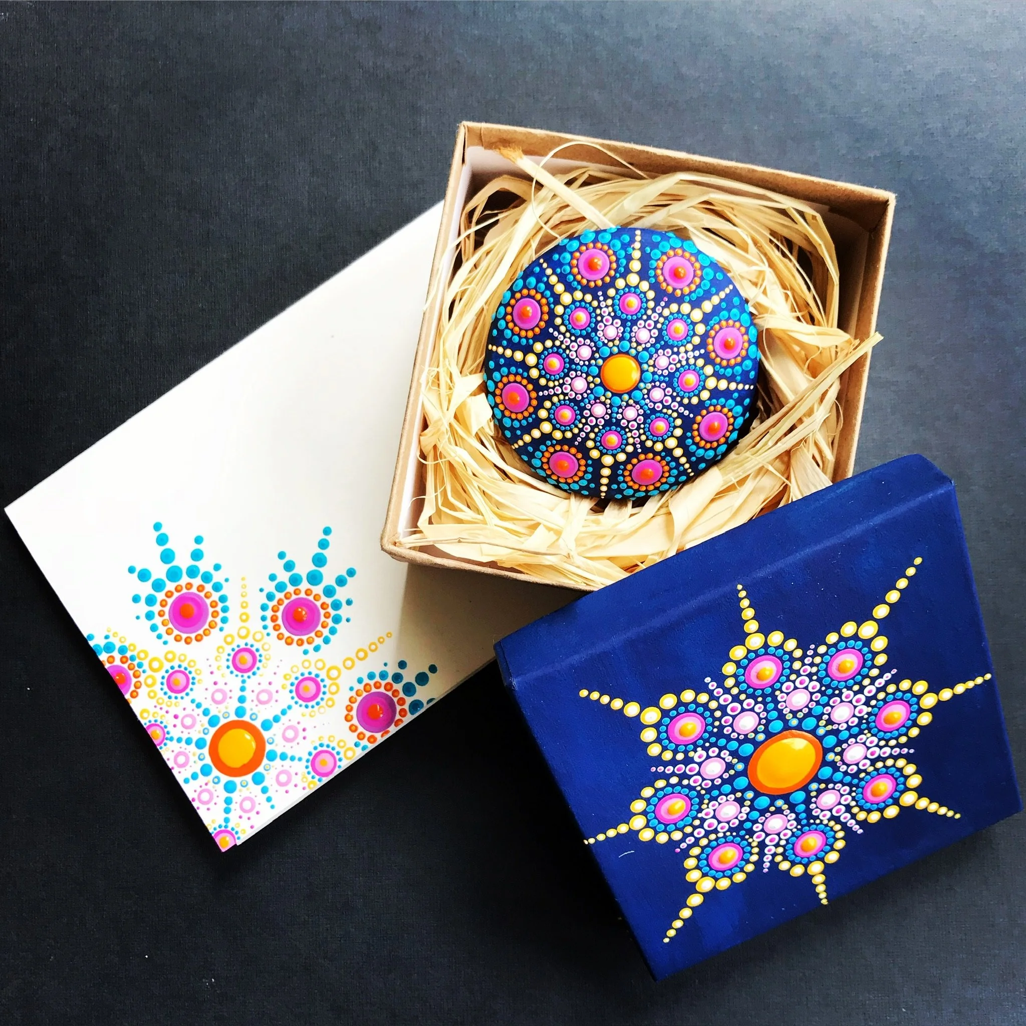 A painted rock with colorful dot patterns in pink, orange, yellow, white, and blue, placed in a small cardboard box with straw filling, on a dark and white background.