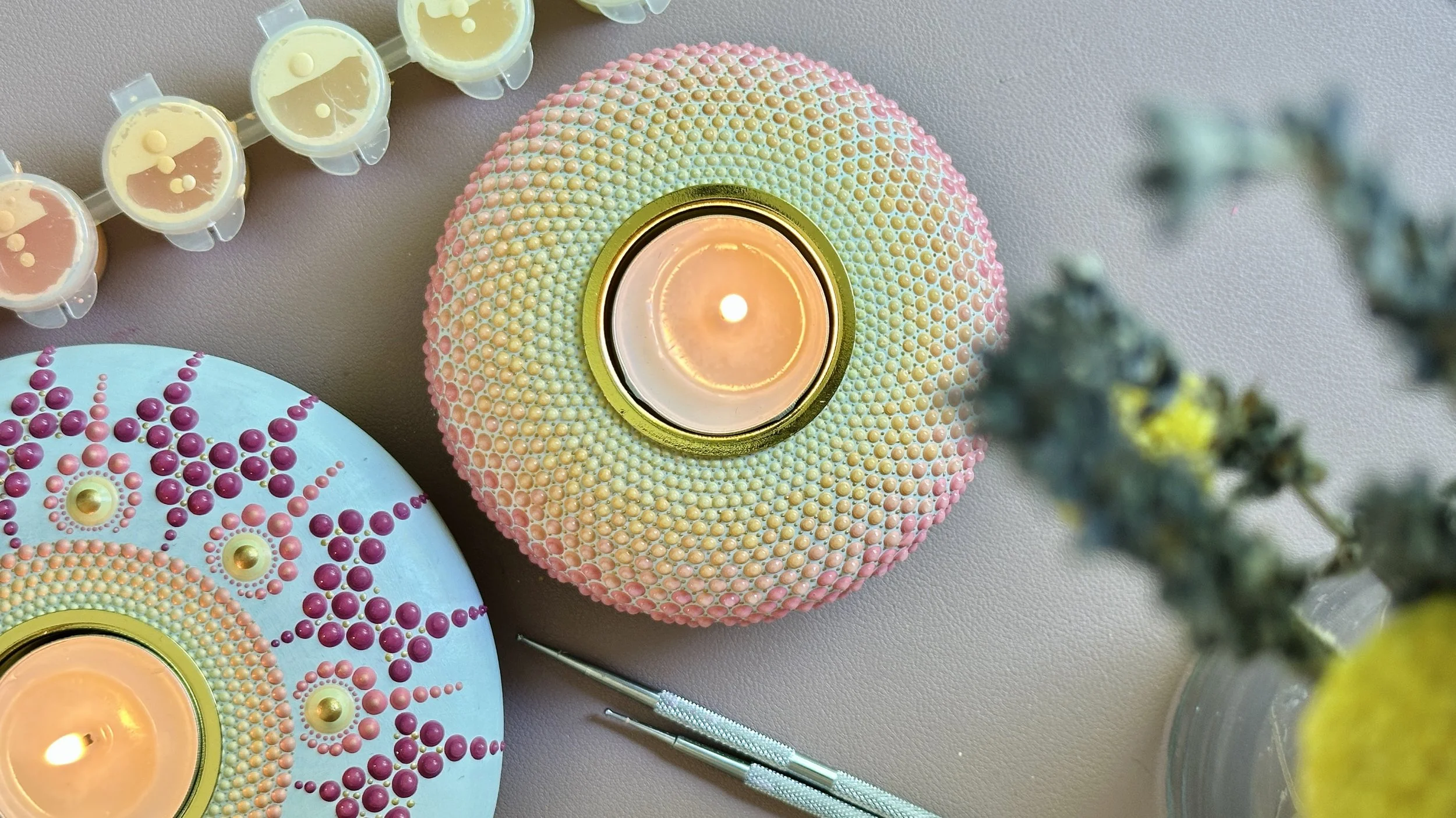 Decorative candles with intricate bead patterns and bright colors, some with lit flames, on a neutral surface next to floral elements.