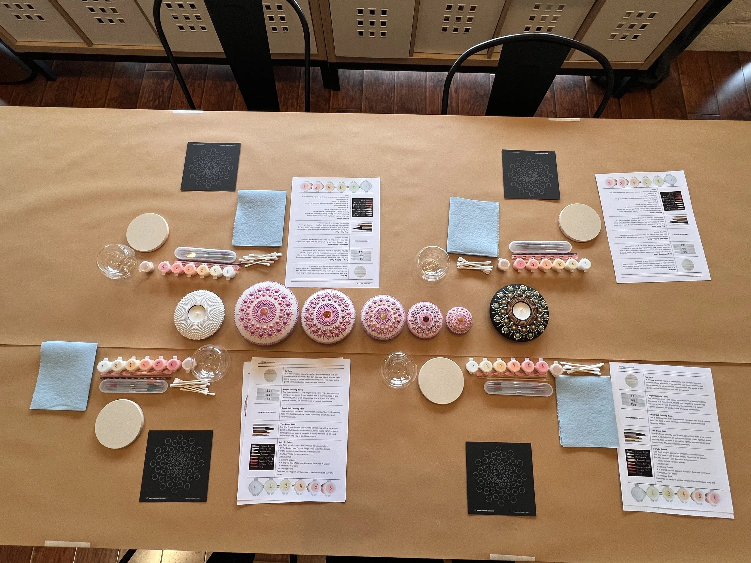Table with art supplies including ceramic paint palettes, paintbrushes, cotton swabs, printed instructions, black templates, paper towels, and small glass bowls, set up for a painting or crafting activity.